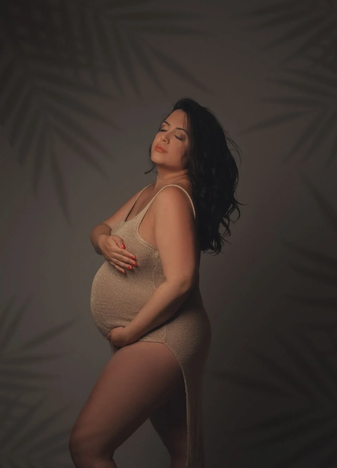 A pregnant woman with long, dark hair posing against a background with palm leaf shadows, wearing a beige dress, with one hand on her belly and the other on her chest, eyes closed.