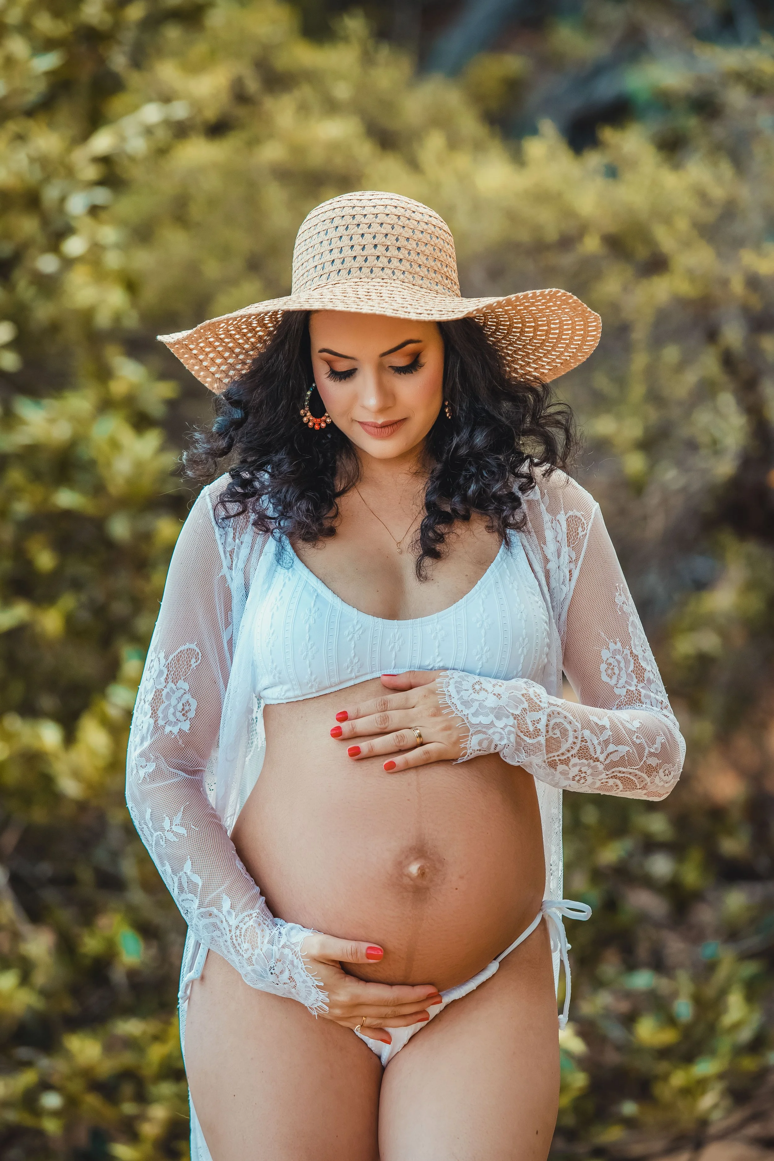 Pregnant woman wearing a wide-brimmed straw hat, floral lace cover-up, and white lingerie, standing outdoors and looking down at her baby bump with a gentle smile.