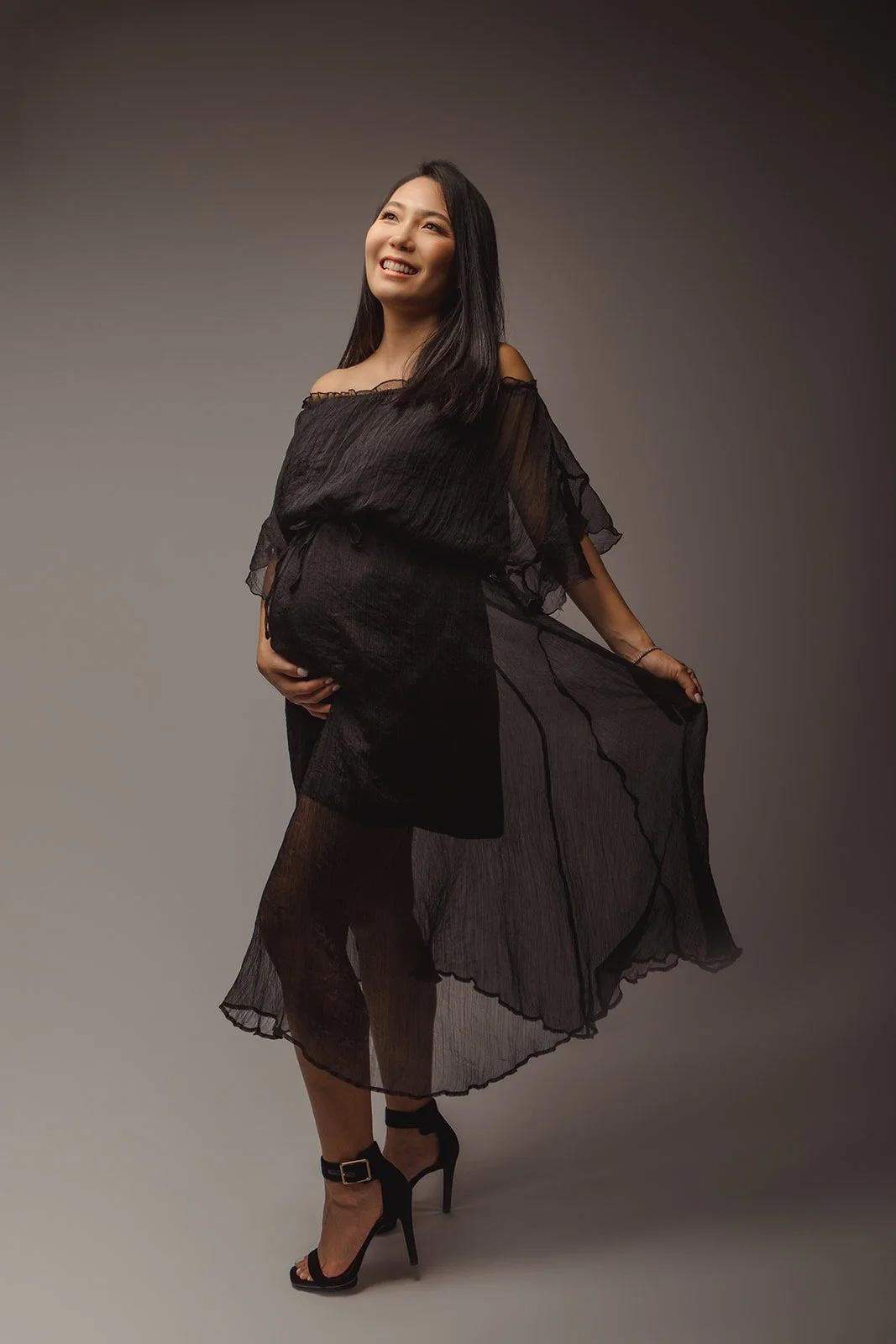 A pregnant woman with long black hair smiling, holding the side of her black, sheer dress, standing against a plain backdrop.