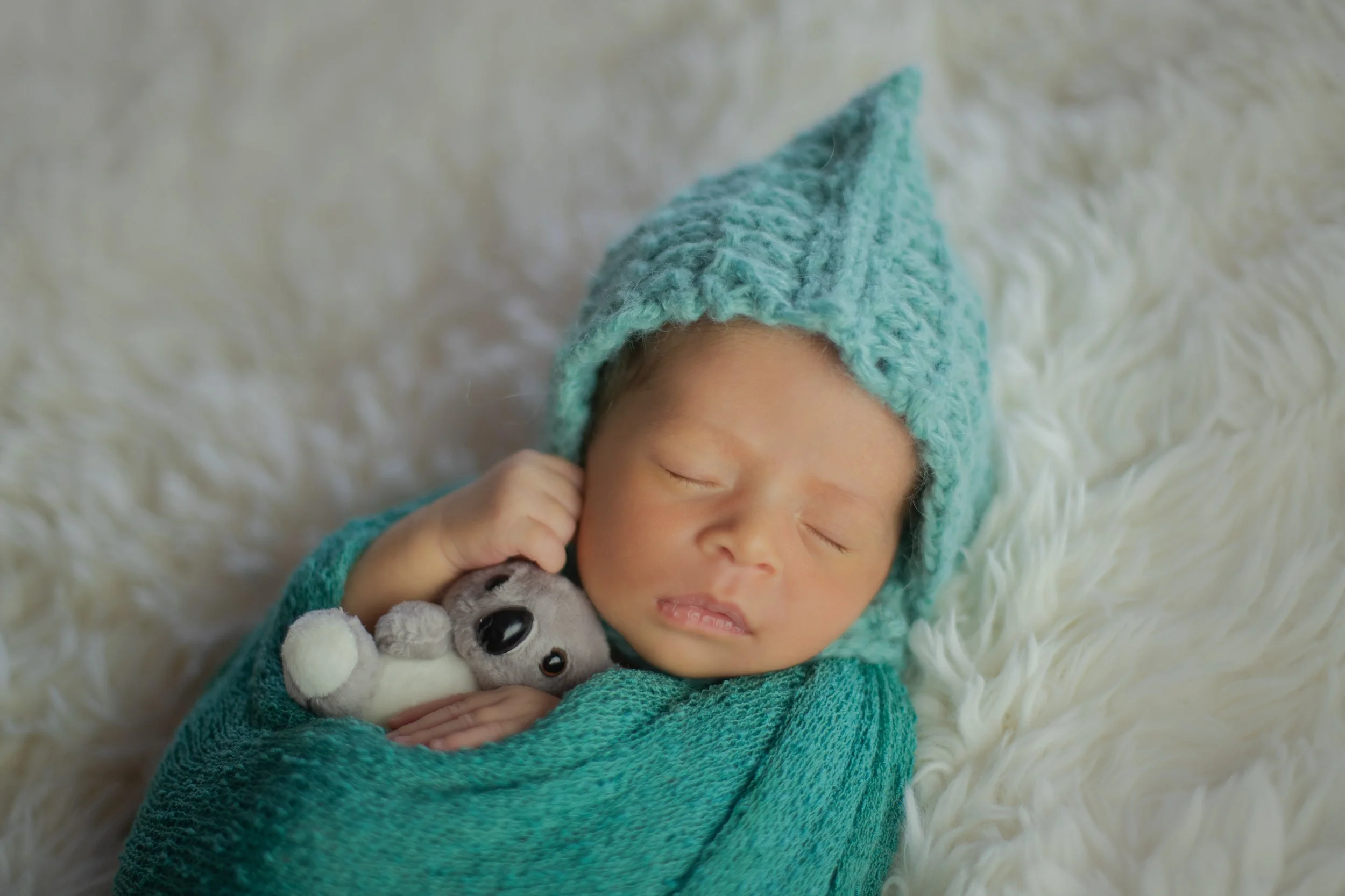 A newborn baby peacefully sleeping on a soft white fur blanket, wearing a blue knitted hat and matching blanket, holding a small grey stuffed animal.