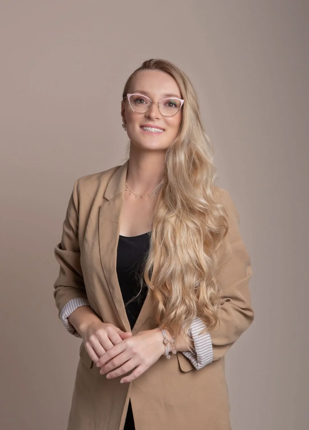 A woman with long wavy blonde hair wearing glasses, a tan blazer over a black top, standing in front of a plain beige background, smiling at the camera.