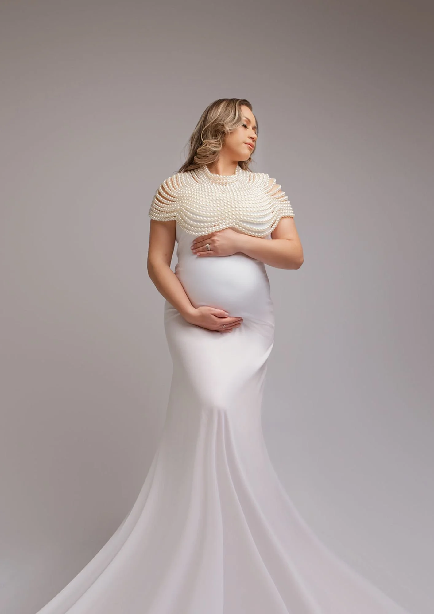 Woman in a white gown with a pearl cape, standing against a plain background, gently cradling her pregnant belly.
