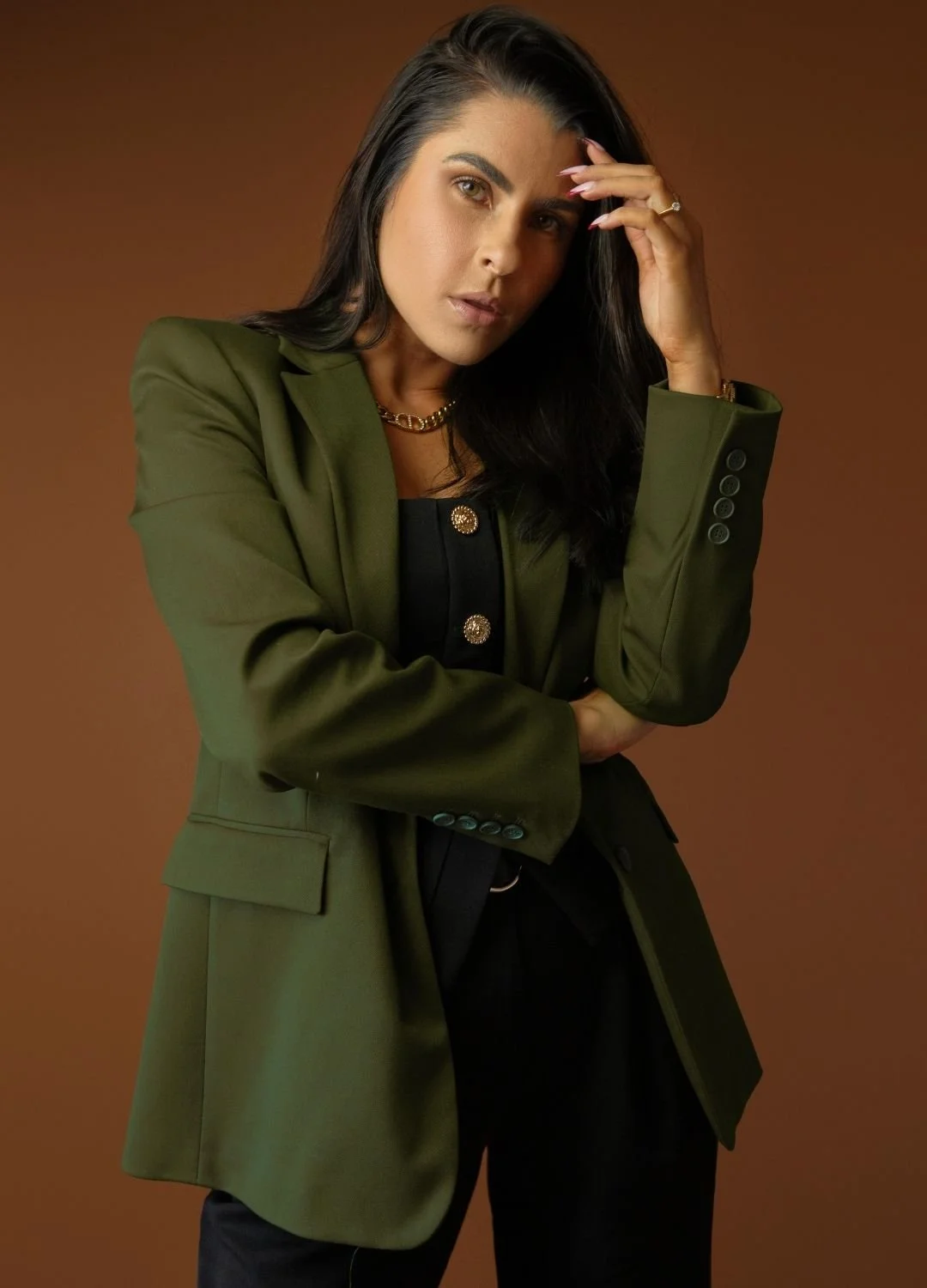 A woman with dark hair, wearing an oversized green blazer, a black top, and black pants, posing with her hand on her forehead against a brown background.