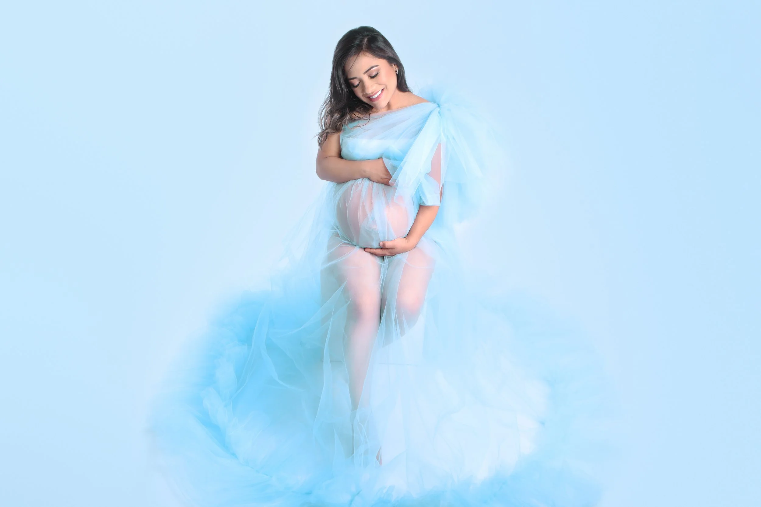 Pregnant woman in a sheer blue dress smiling and holding her belly, standing against a light blue background.