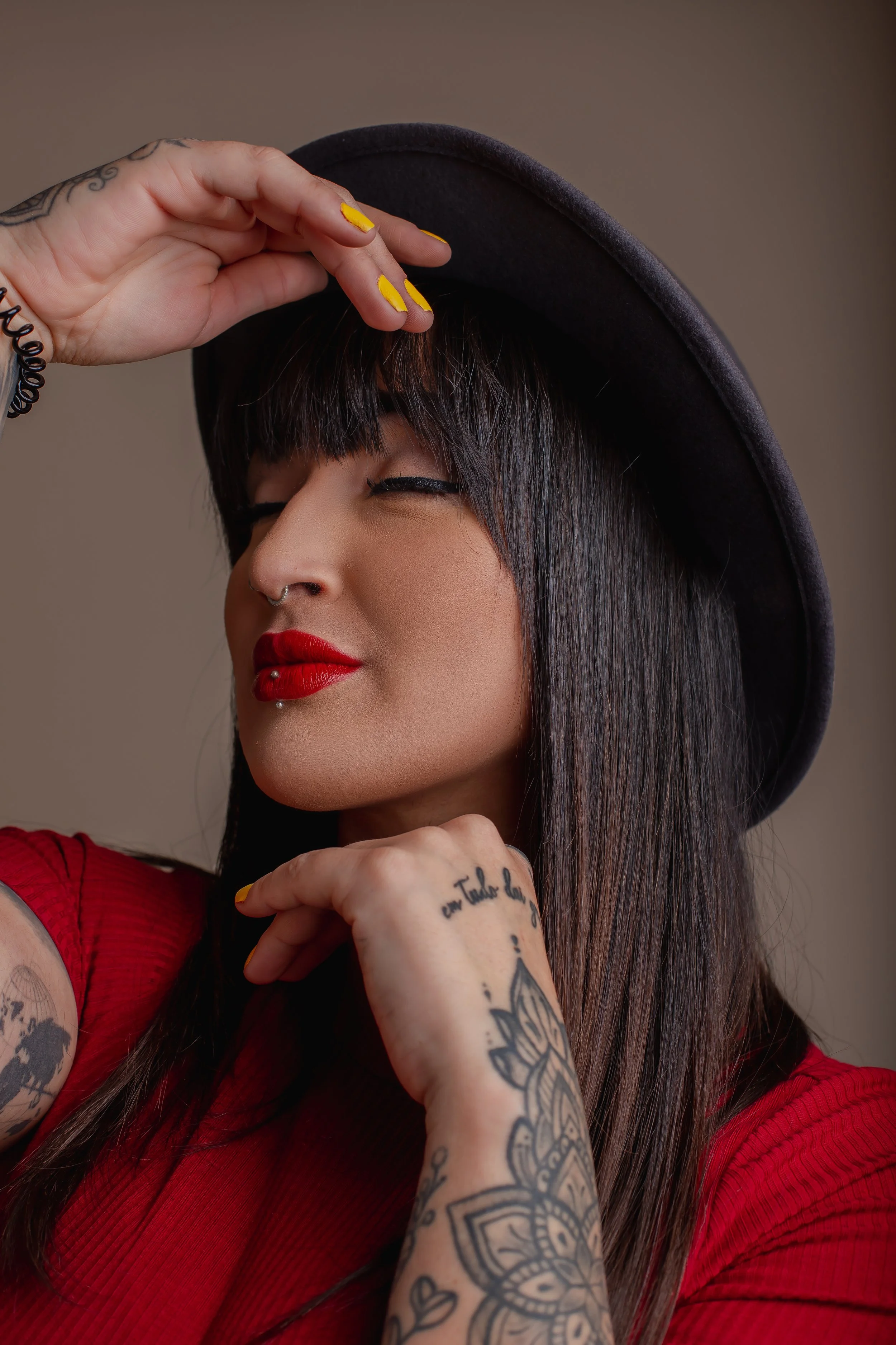 A woman with long dark hair, red lipstick, tattoos on her arm, and a nose piercing wears a red top and a black hat. She is smiling with eyes closed, touching her hat with one hand and resting her chin on her other hand.