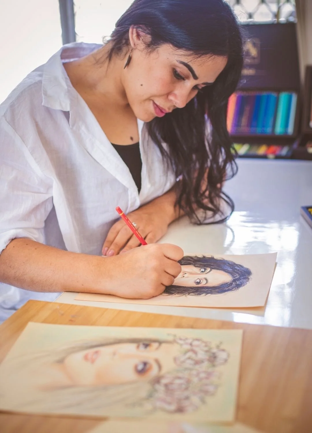 A woman with dark hair and wearing a white shirt is drawing a portrait of a woman with glasses and long dark hair, using colored pencils on a piece of paper, sitting at a wooden table.