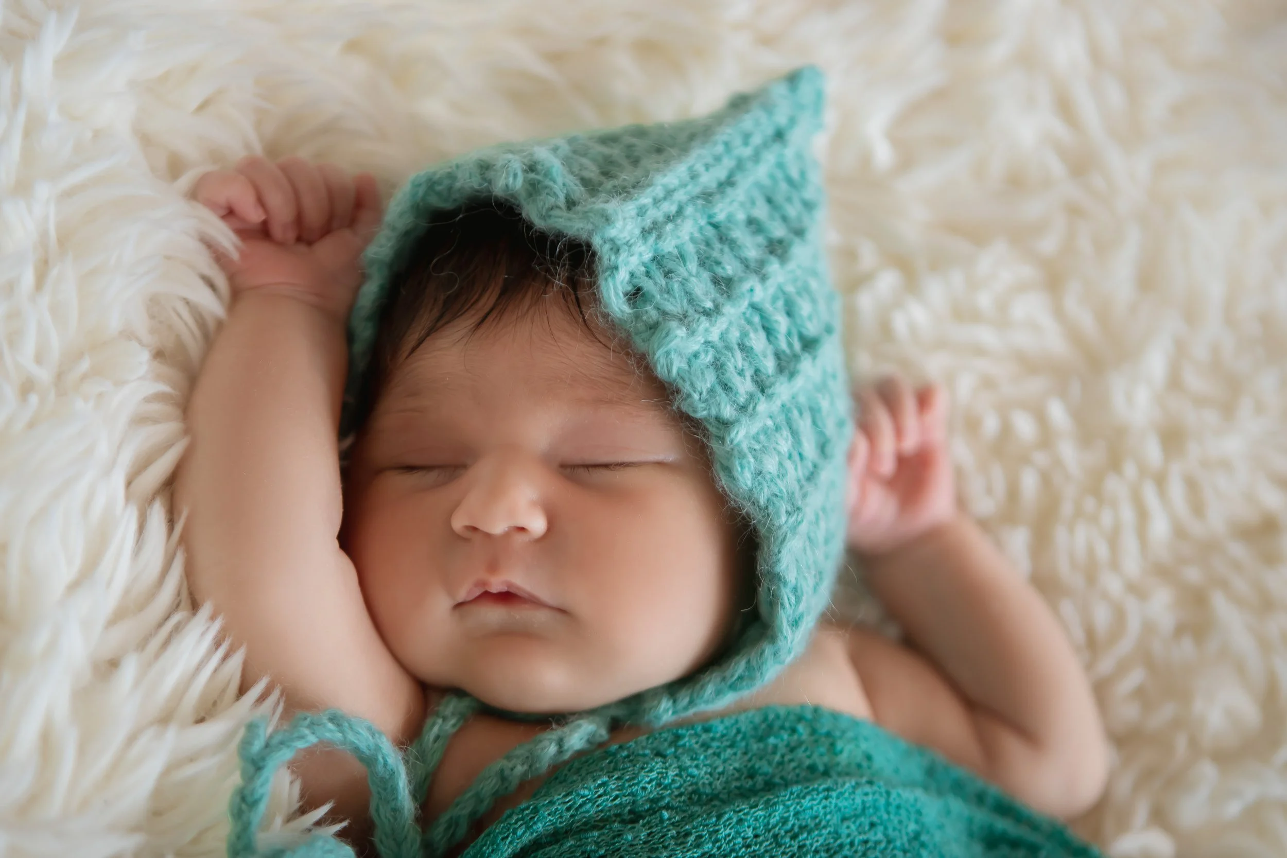A sleeping baby with a turquoise knit hat and matching wrap, resting on a fluffy white surface.
