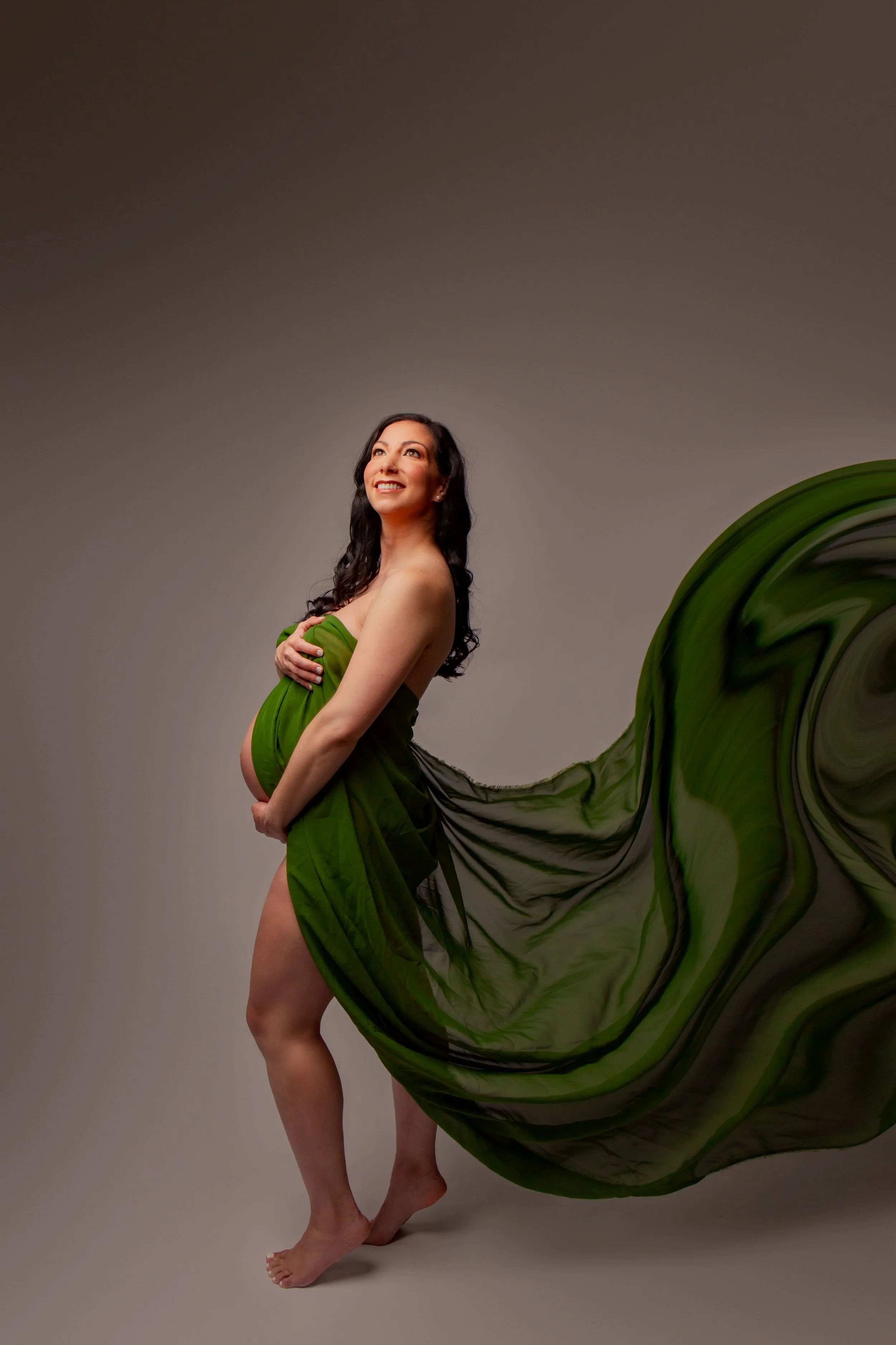 Pregnant woman with long dark hair, smiling, partially wrapped in green fabric, standing barefoot with a flowy green cloth extending behind her, inside a plain studio.