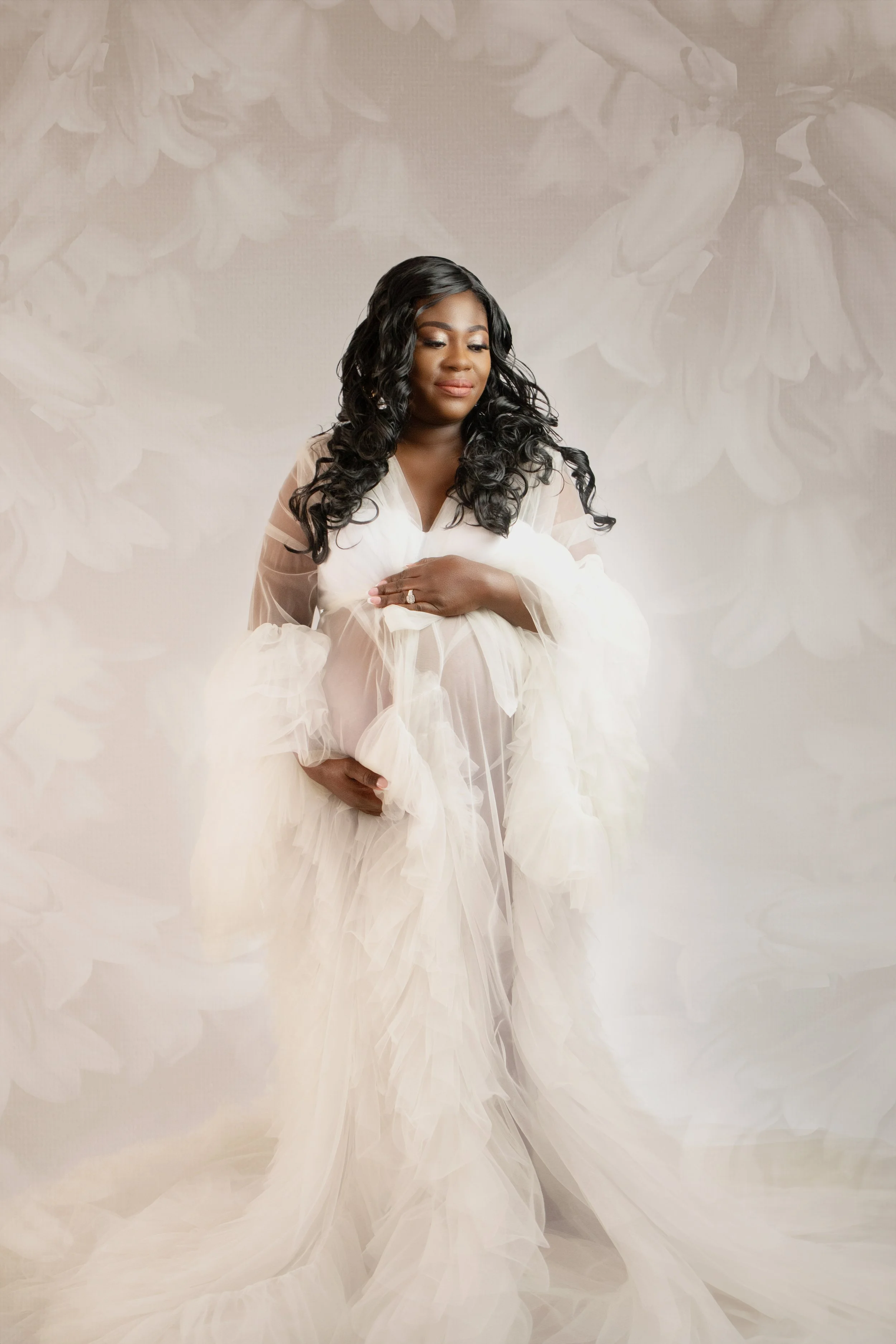 A woman dressed in a flowing white gown, holding her pregnant belly, standing against a soft, floral-patterned background.