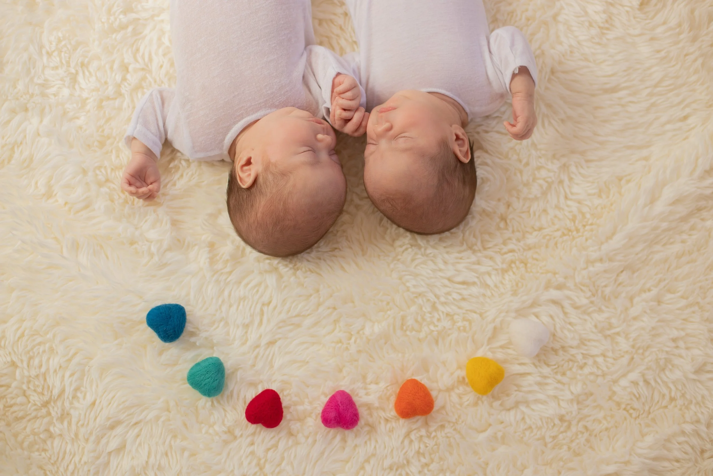 Two newborn babies lying on a soft, cream-colored blanket, facing each other with their heads touching, colorful felt hearts arranged in a semi-circle at the bottom of the image.