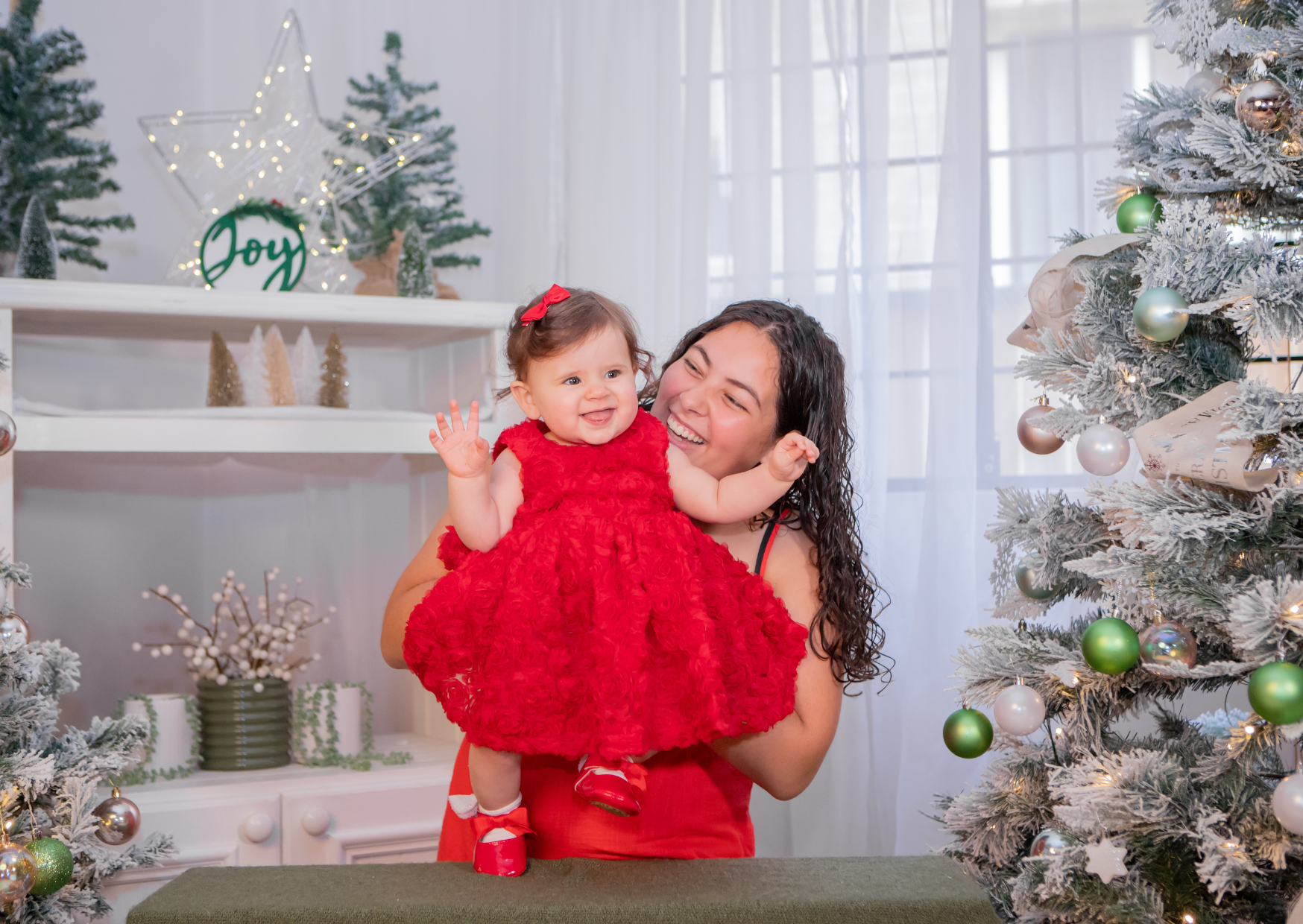 A woman and a young girl in a red dress smiling and playing around Christmas decor, including a Christmas tree, ornaments, and festive decorations in a brightly lit room.