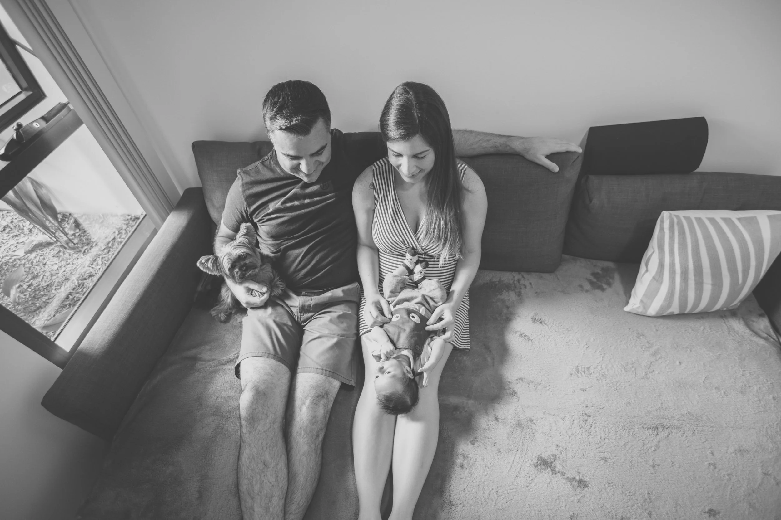 A family of three sitting on a couch with their dog, all looking at a child lying on the woman's lap, in a cozy living room.
