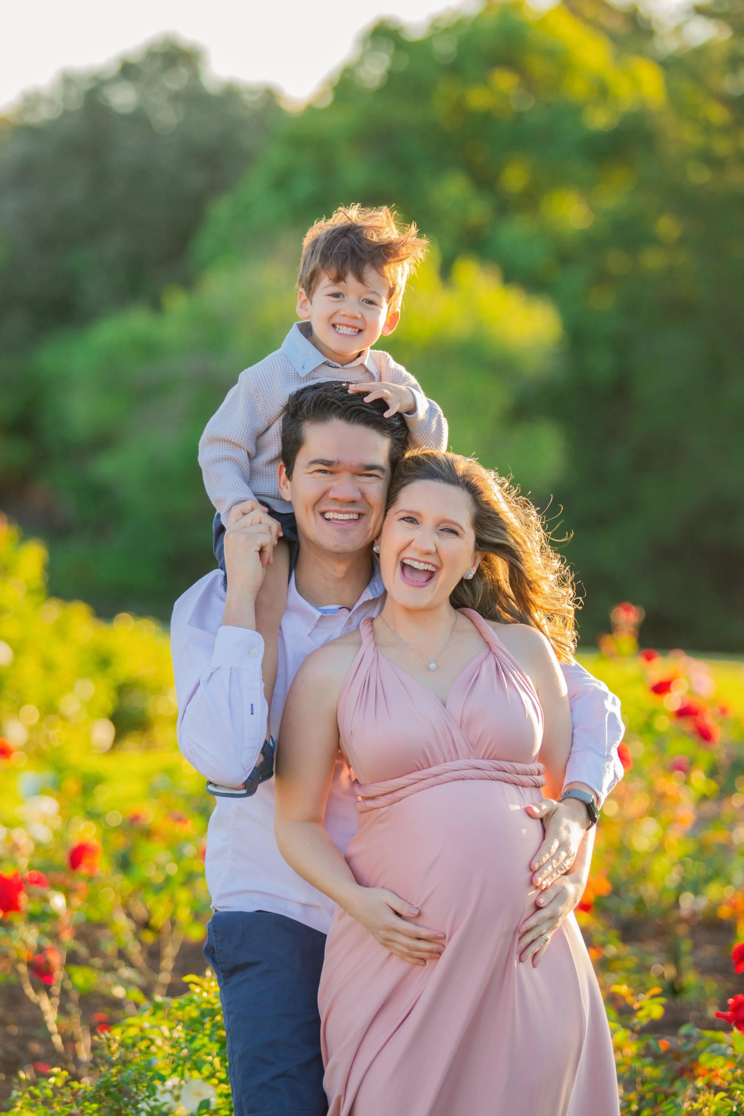 A happy family of three, including a pregnant woman, a man, and a young boy, smiling outdoors in a garden with colorful flowers during the daytime.