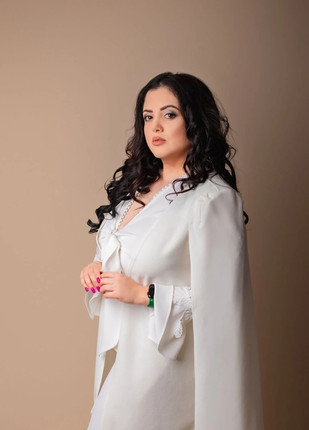 A woman with dark curly hair wearing a white dress and cape, standing against a beige background, looking at the camera.