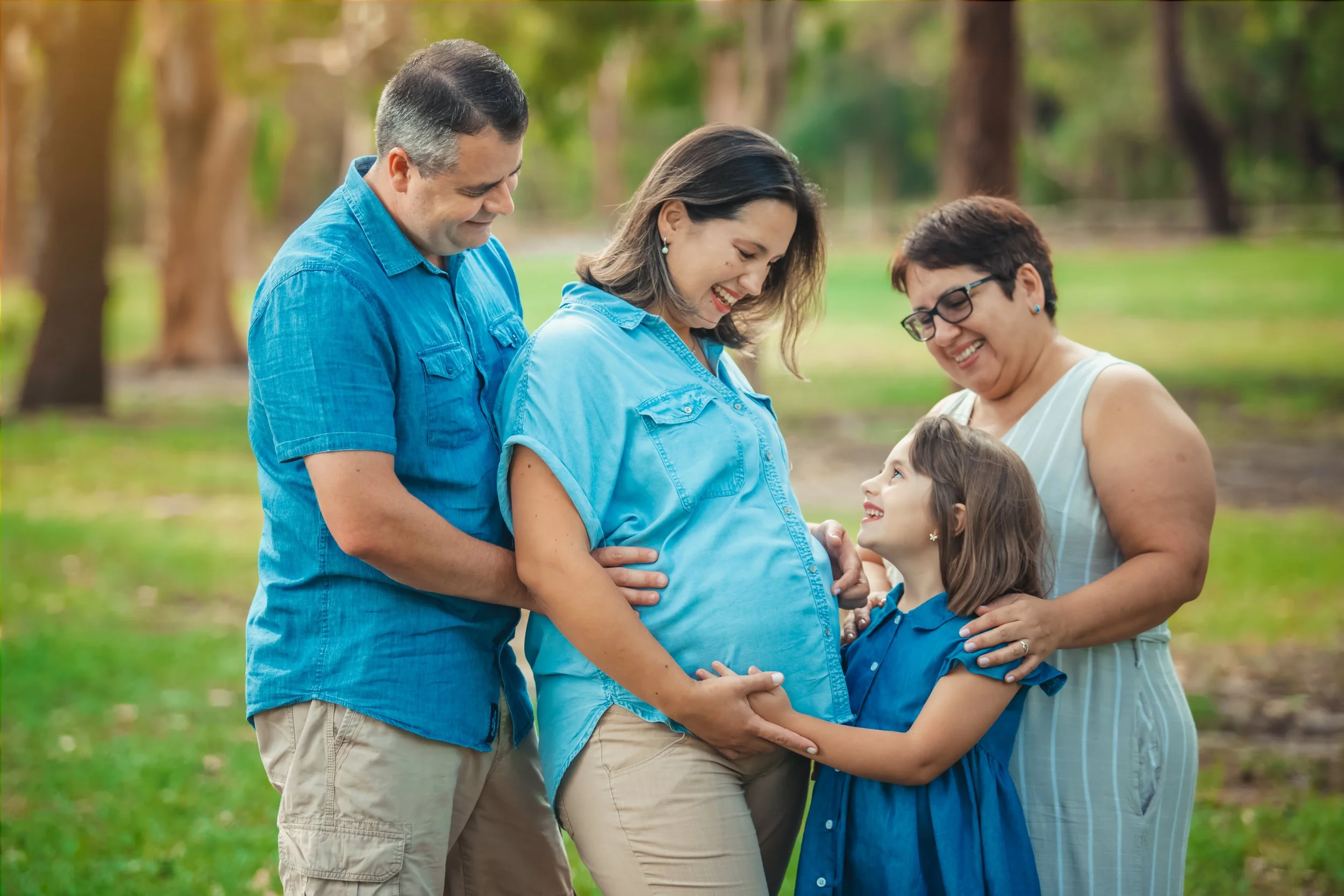 A pregnant woman smiling at a young girl touching her belly, surrounded by three adults in a park with trees, all sharing a joyful moment.