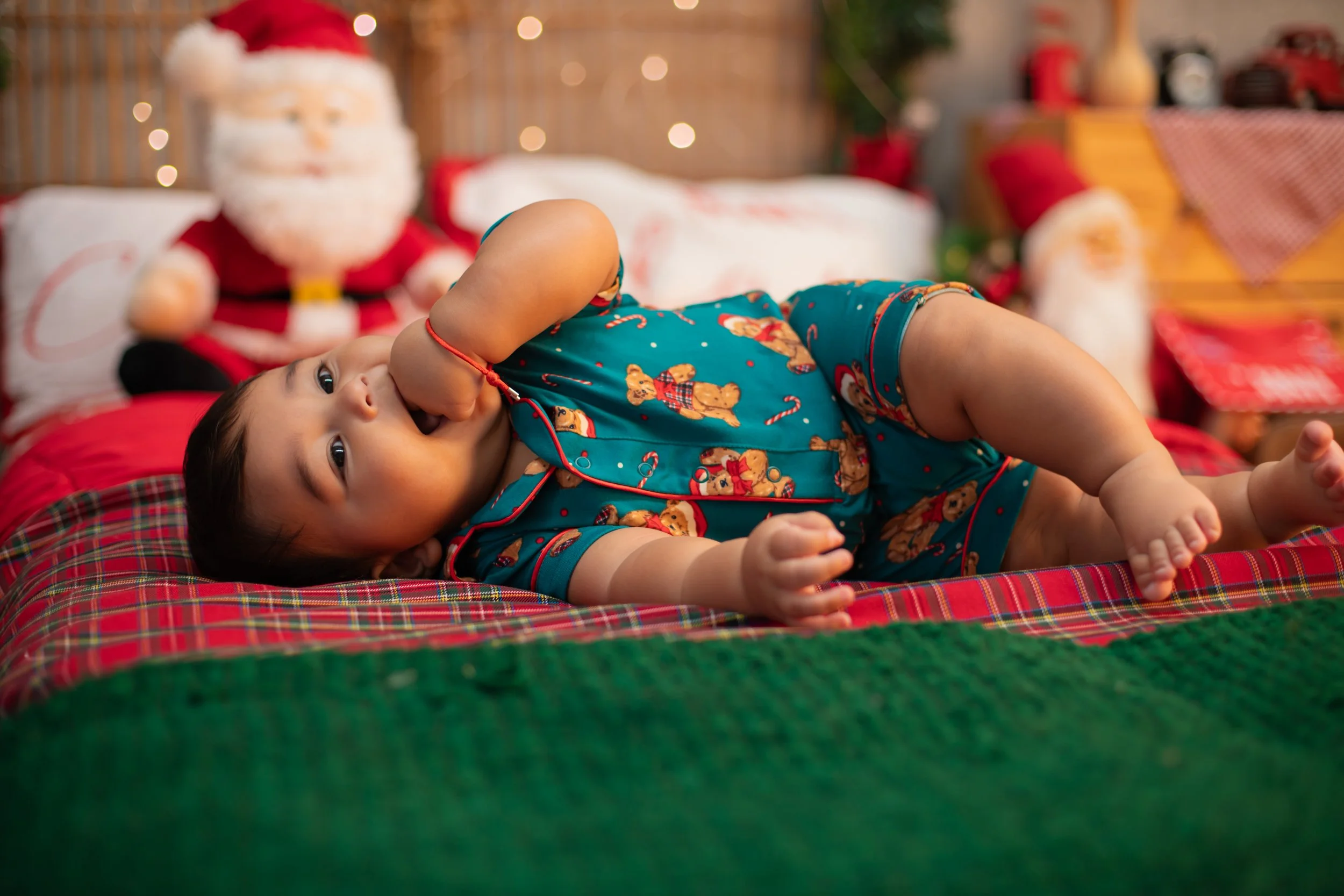 A young child lying on a bed with Christmas-themed pajamas, holding a finger in her mouth. The bed has red and green blankets, and a Santa Claus plush toy and Christmas decorations are visible in the background.