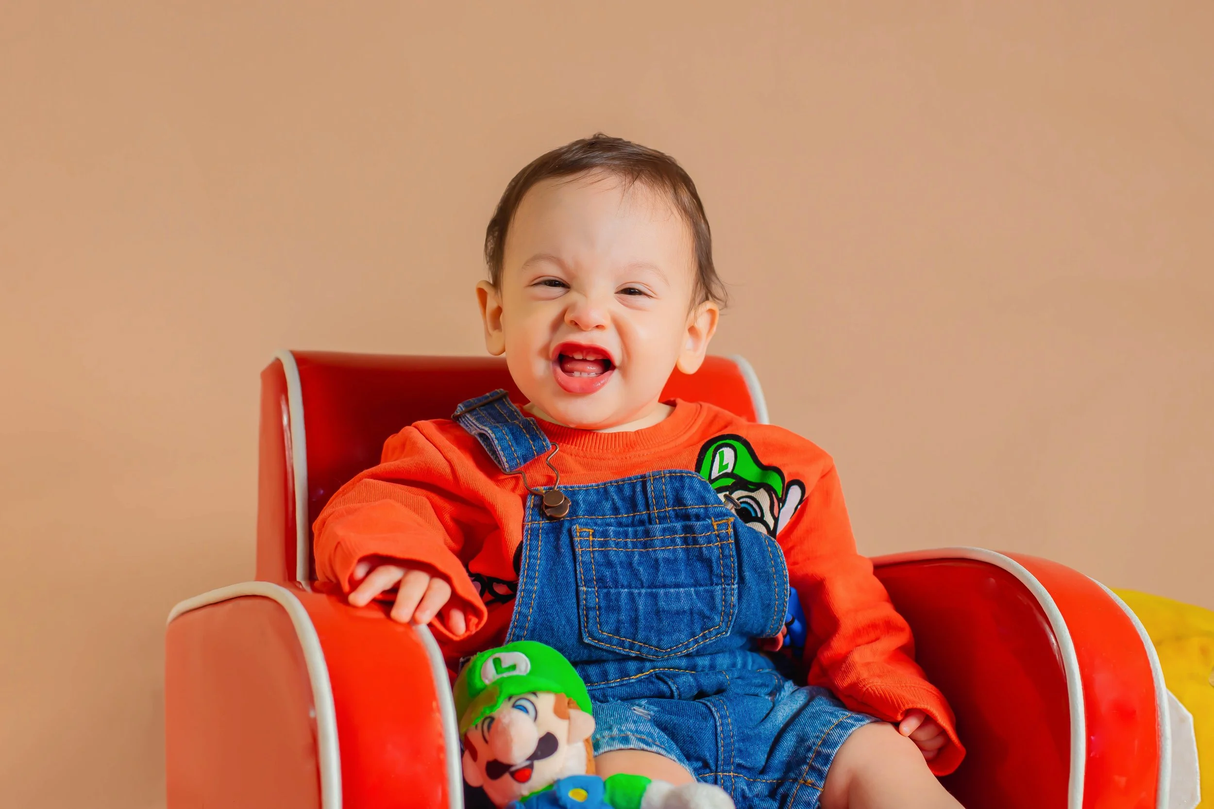 Smiling toddler dressed in Mario-themed clothes sitting in a red chair with a Luigi plush toy, against a beige background.