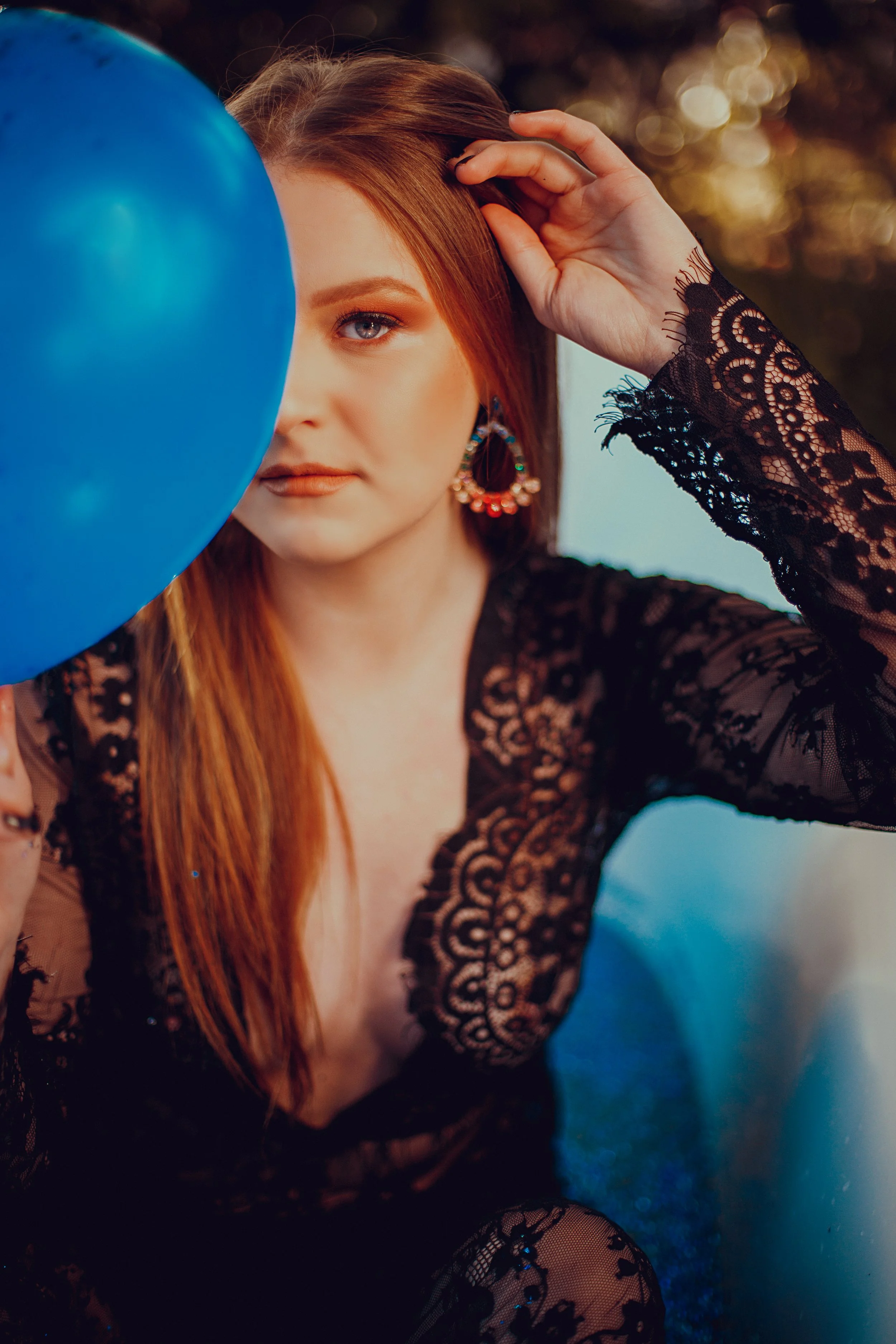 A woman with red hair and lace dress holding a blue balloon, partially blocking her face, outdoors during sunset.