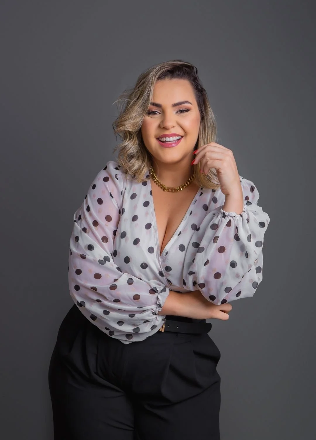 A woman with blonde, shoulder-length hair styled in loose waves, smiling and making eye contact with the camera. She's wearing a white blouse with black polka dots, a gold chain necklace, and black pants, standing against a plain gray background.