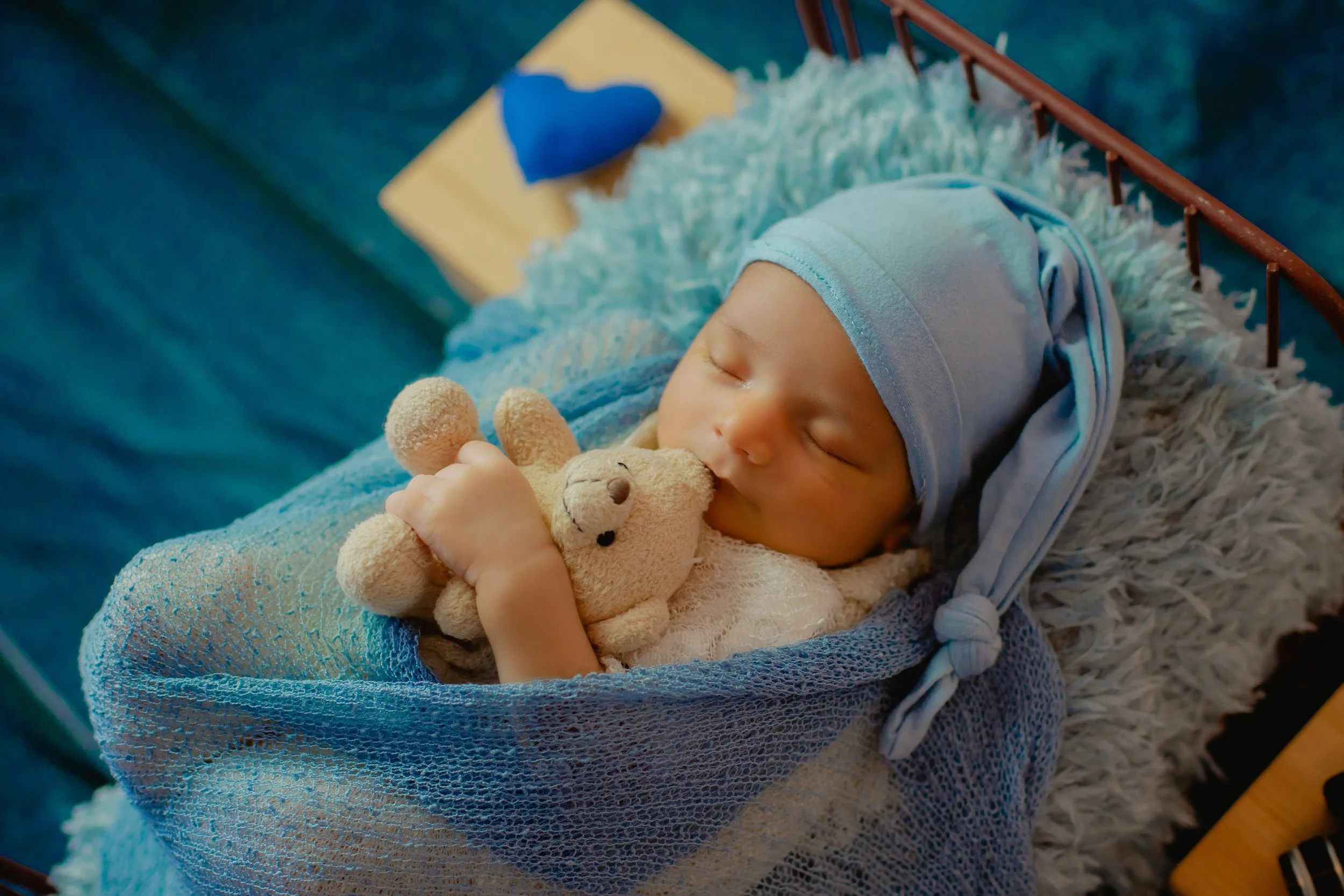 A sleeping baby wearing a blue hat lies in a crib, hugging a small stuffed bear.