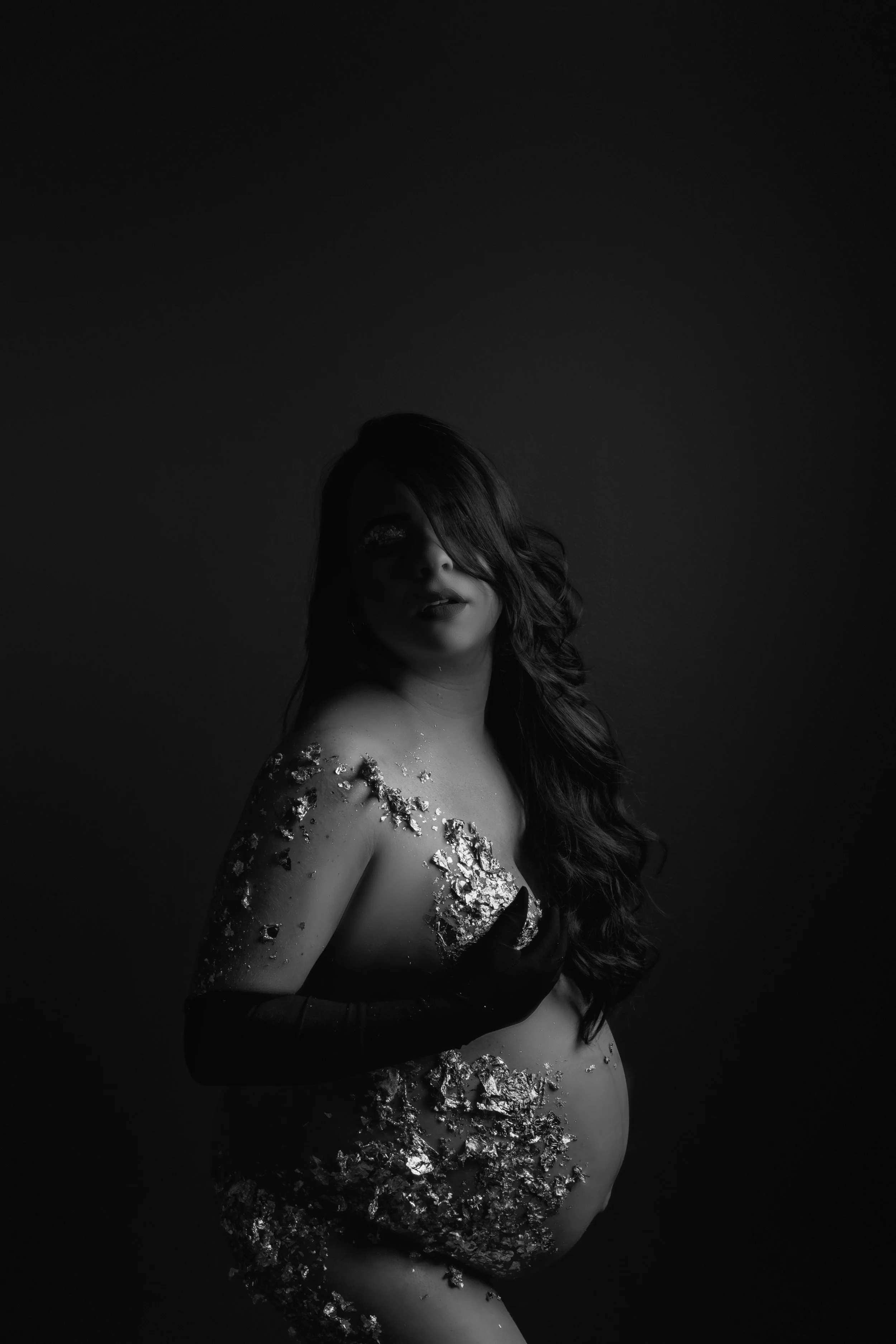 Black and white photo of a woman with long hair, holding her chest, covered in gold flakes, with a dark background.