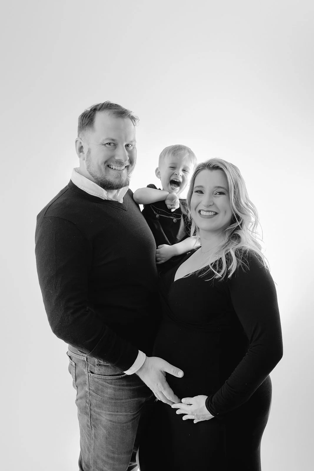 A happy family portrait of a man, woman, and a young child, smiling and laughing, with the woman pregnant, in black and white.