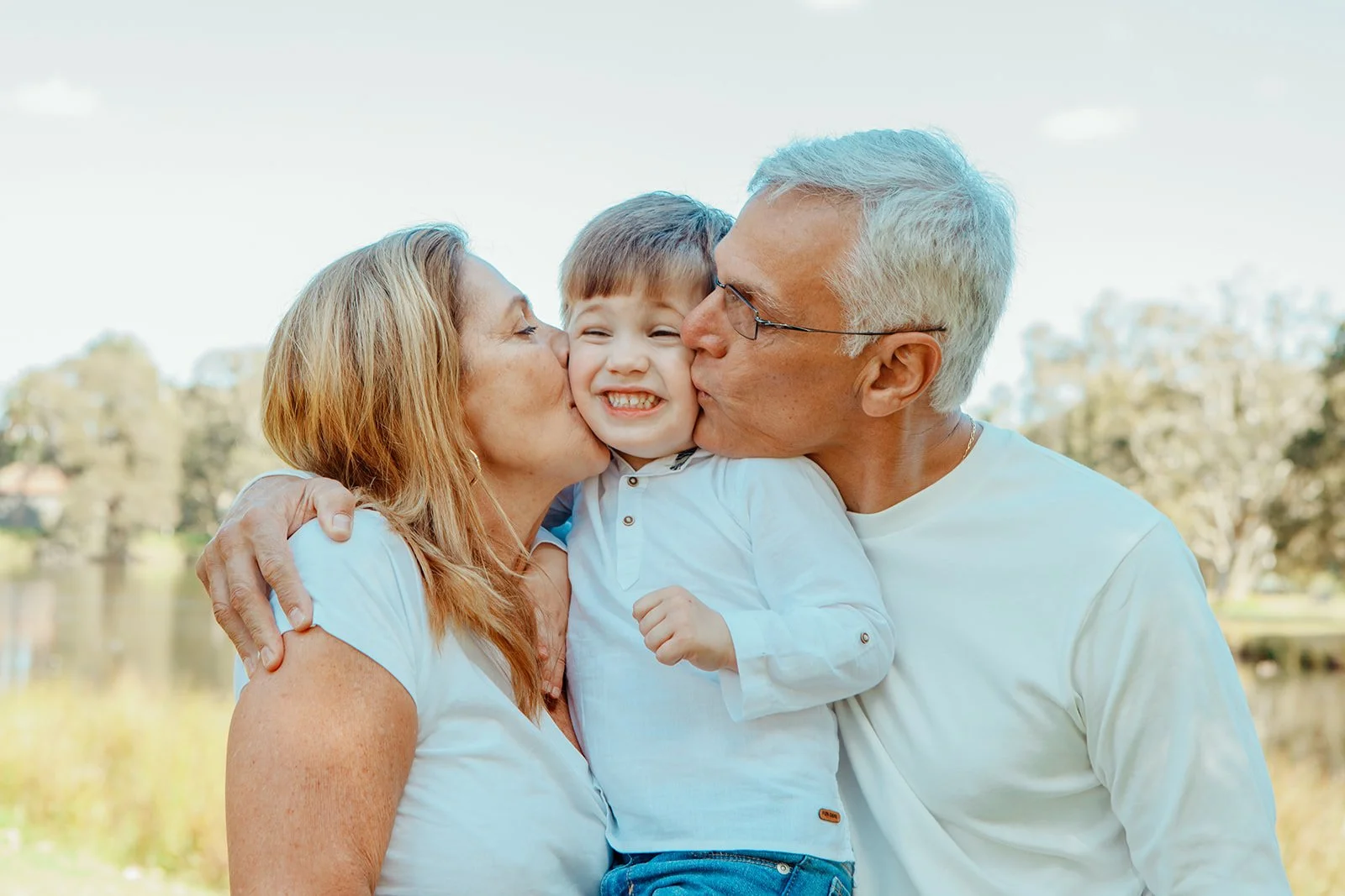Smiling young boy being kissed on each cheek by a woman and an older man outdoors by a lake with trees in the background.