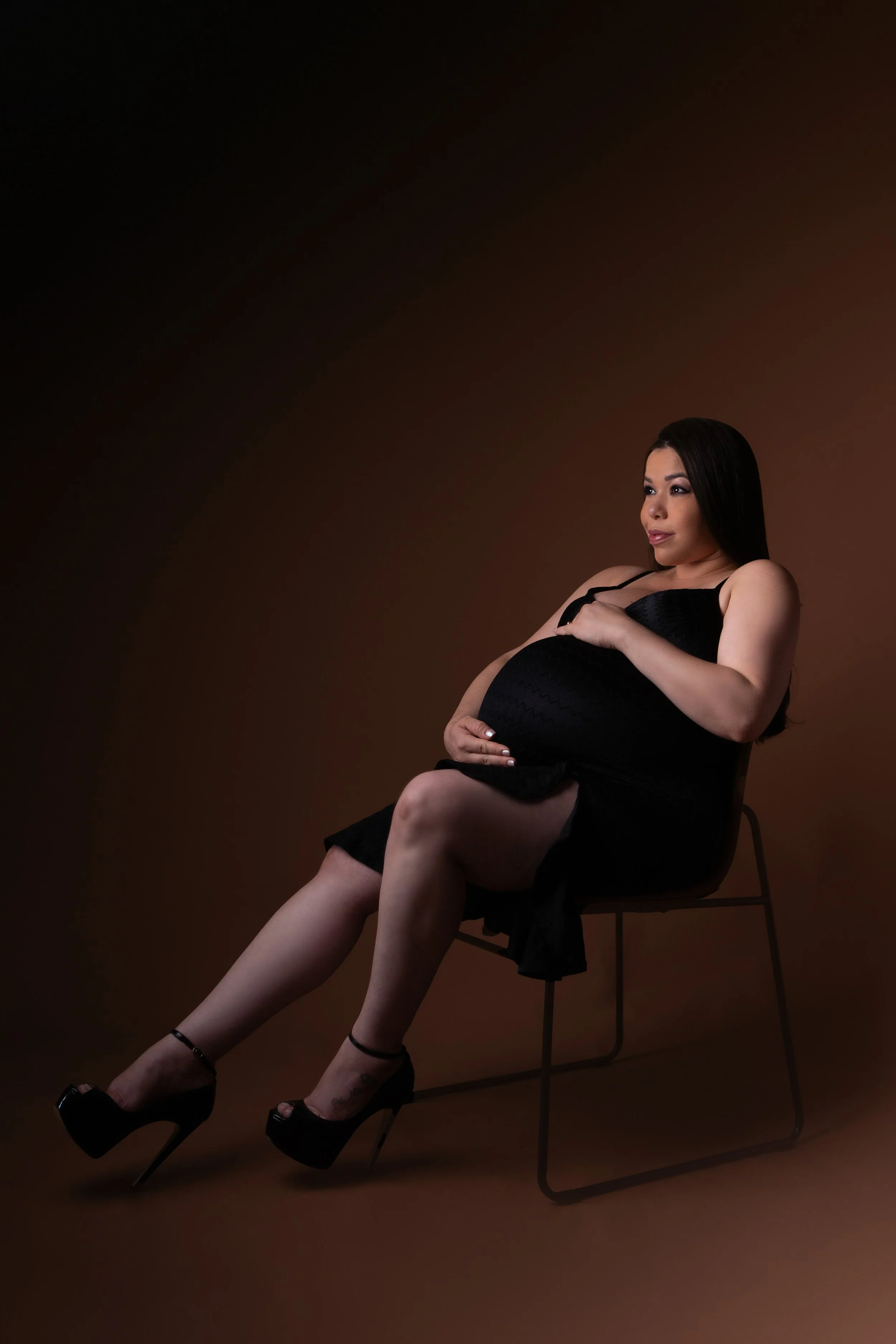 Maternity portrait of a pregnant woman sitting on a chair, wearing a black dress and high heels, holding her belly with her right hand, against a dark background.