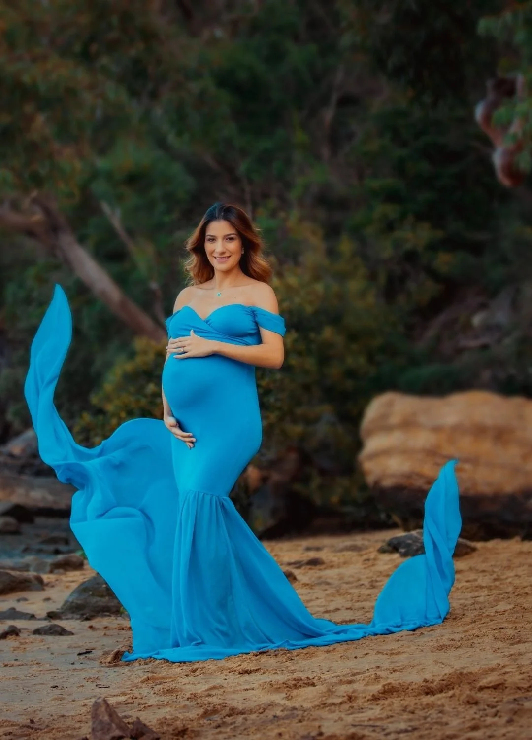 Pregnant woman in a flowing blue dress standing on the beach with a background of trees and rocks.