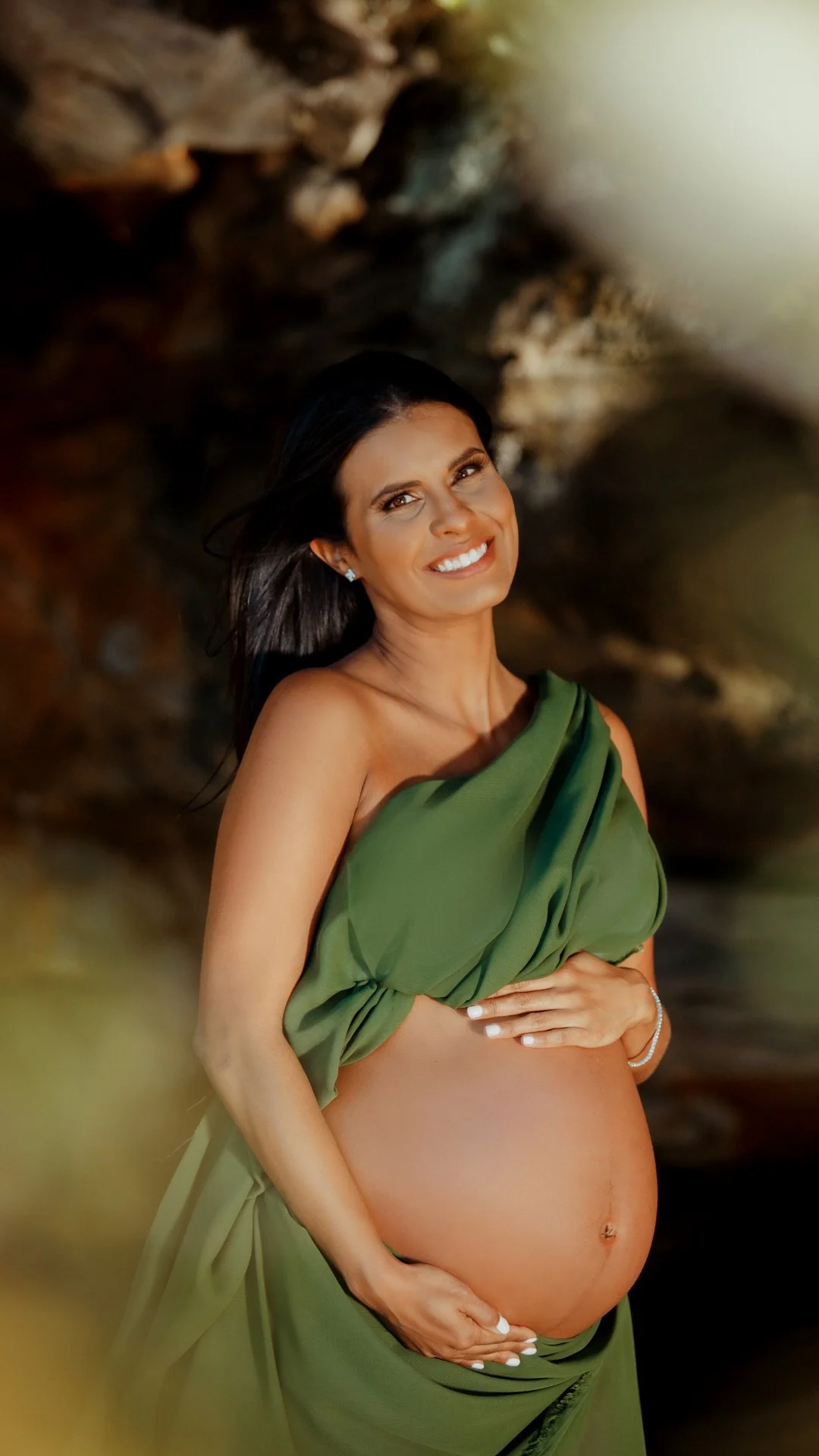 A pregnant woman smiling, wearing a green draped fabric, standing outdoors in front of a blurred rocky background.