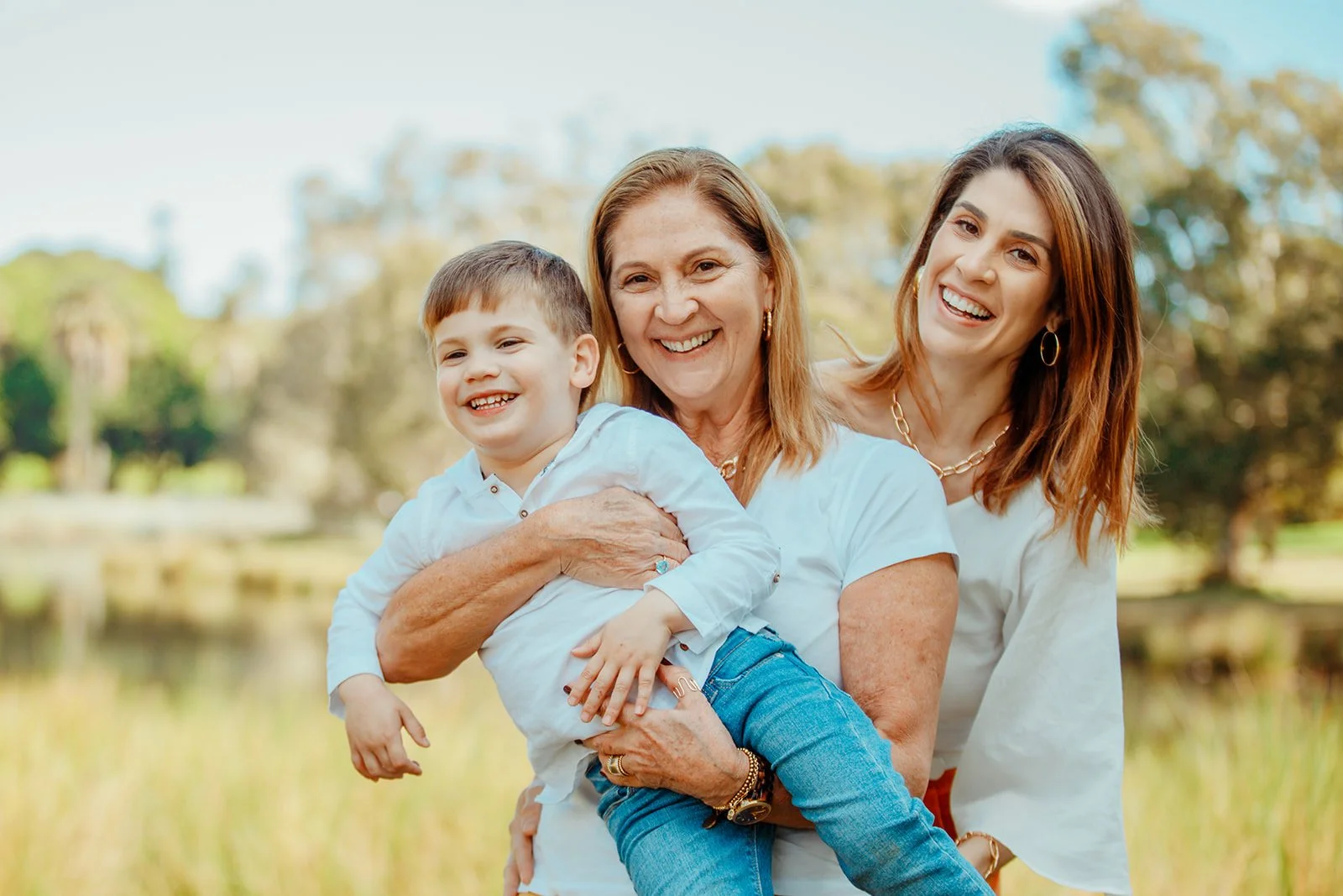Three generations of women smiling outdoors, with a small boy being held by the older woman, in a natural setting with trees and grass.