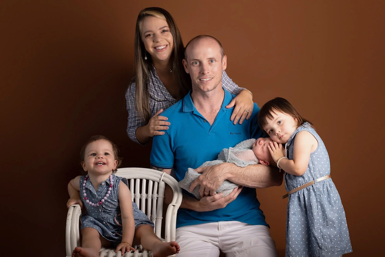 Family portrait with father holding newborn baby, mother standing behind, and two young girls, one sitting and one cuddling the baby, all wearing blue and gray dresses against a brown background.
