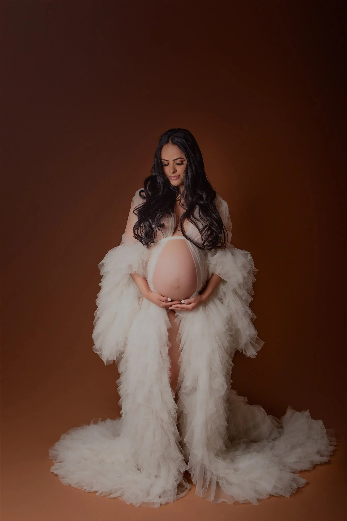 A pregnant woman with long black hair wearing a sheer, flowing, ruffled beige dress, cradling her belly, standing against a brown background.