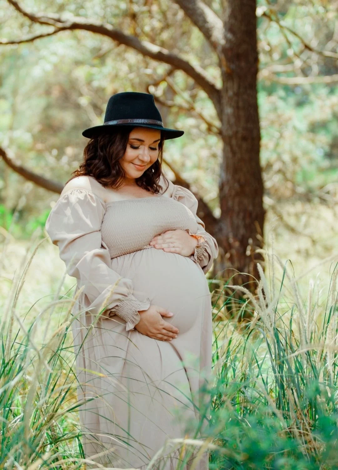 Pregnant woman in a beige dress and black hat standing in a grassy outdoor area with trees, gently holding her belly and smiling.