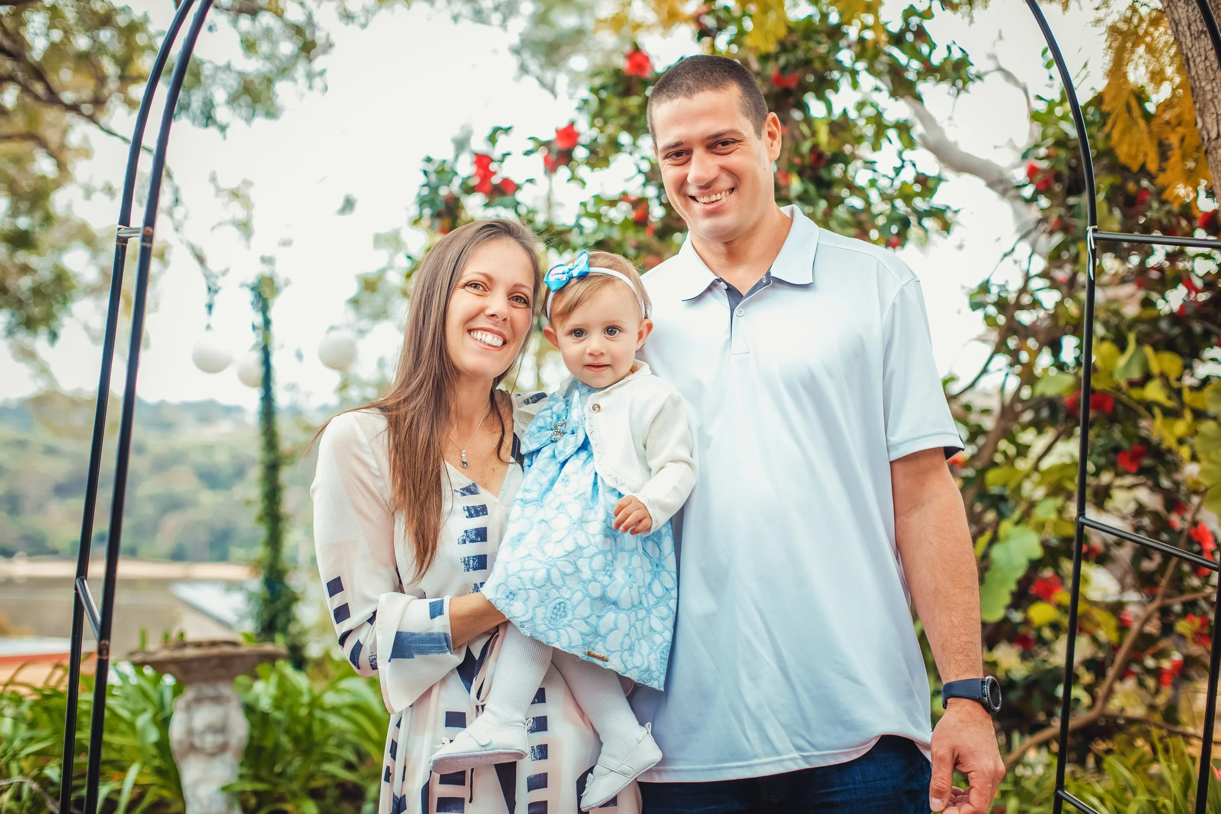 Family of three outdoor portrait: smiling woman holding young girl, man beside them, decorative garden background.