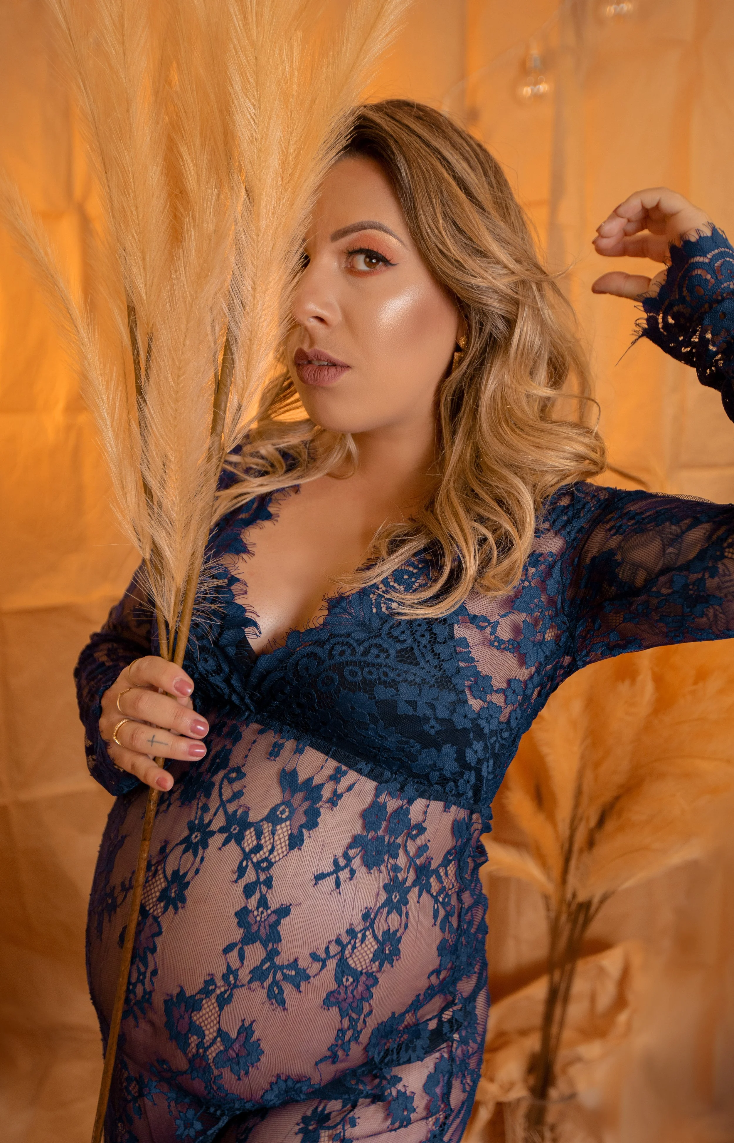 Woman in navy blue lace dress holding pampas grass in front of orange background.