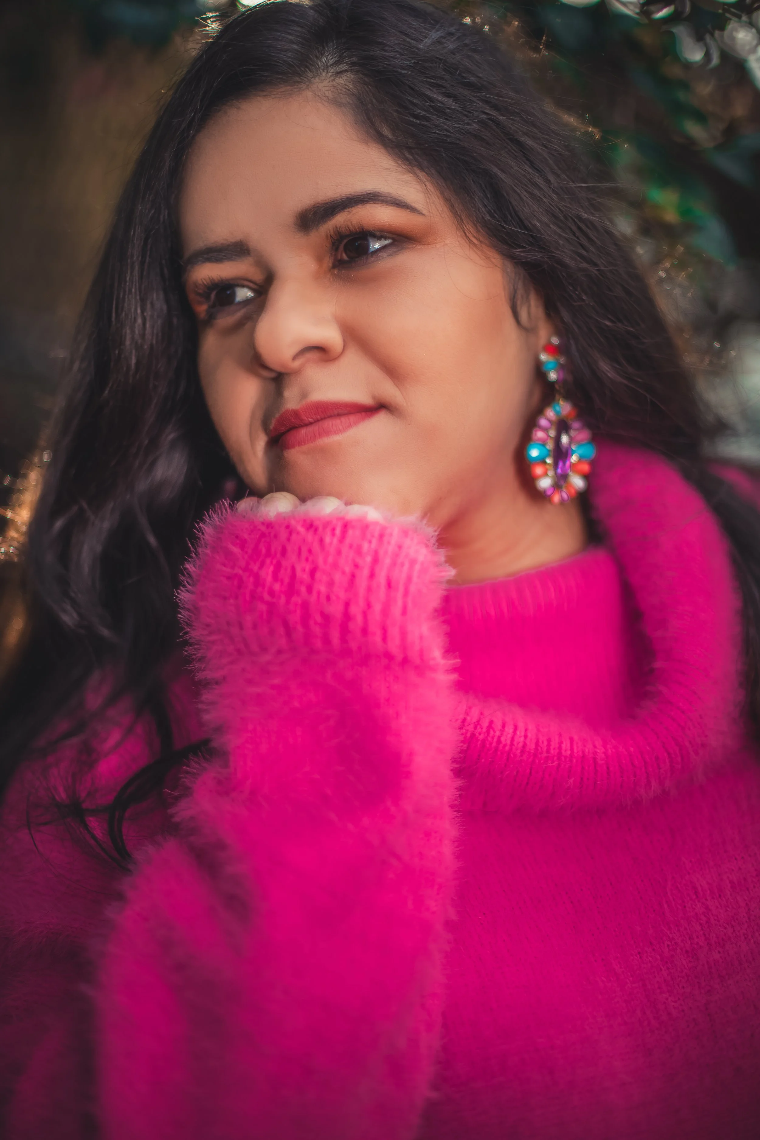 A woman with long dark hair wearing a pink fuzzy sweater and colorful earrings, smiling gently with her chin resting on her hand.