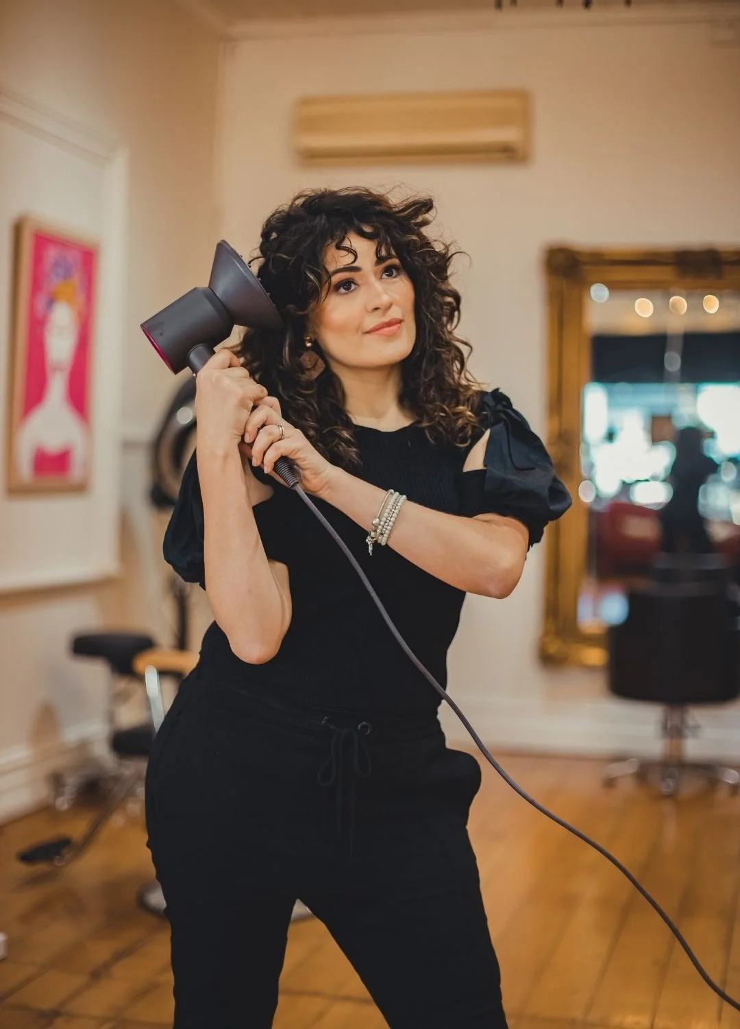 Woman with curly hair holding a black hairdryer, standing in a room with wooden floor and large mirror.