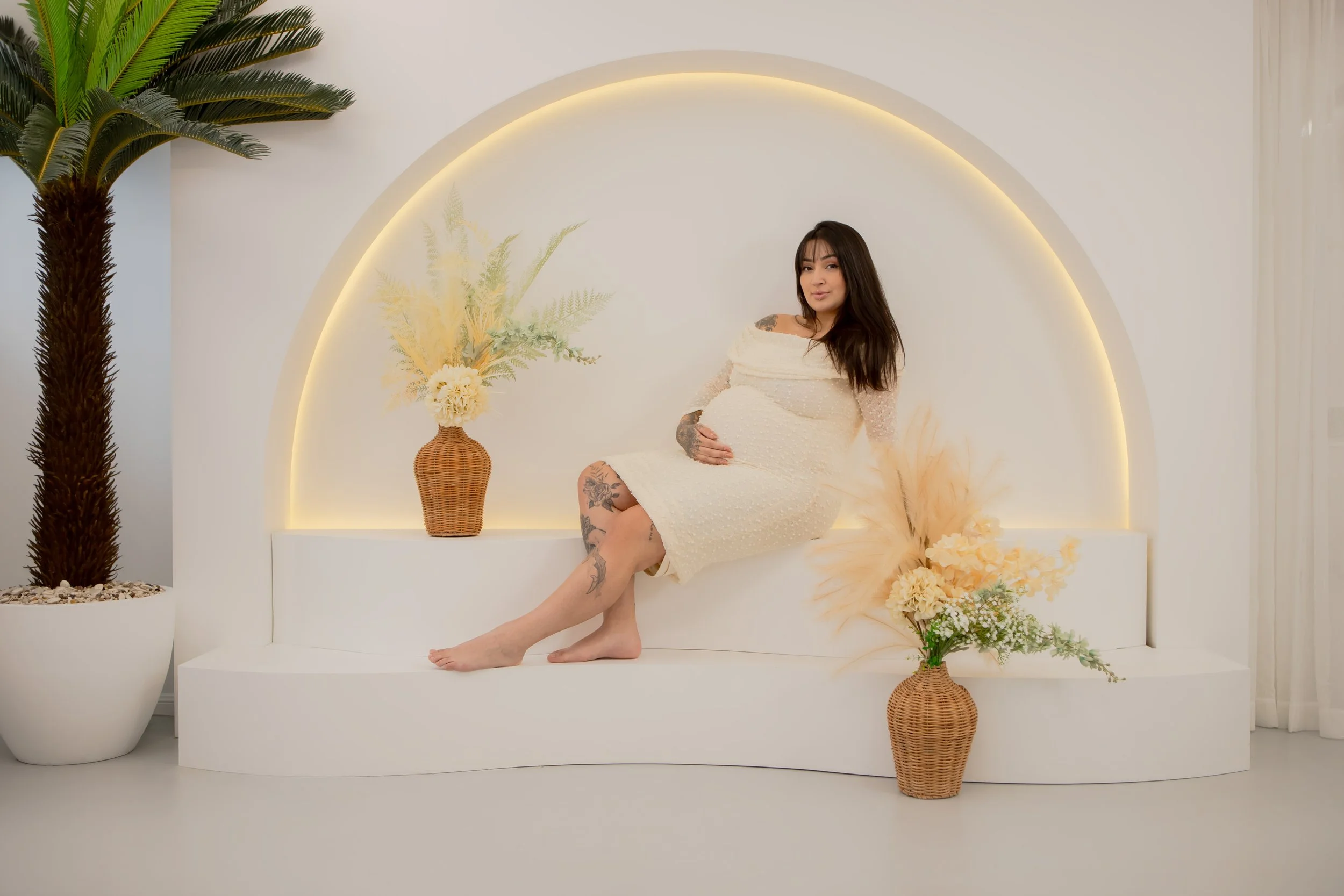 Pregnant woman with dark hair and tattoos sitting on a white ledge in a modern, minimalistic room with a white background, large potted plant, and vases with flowers.