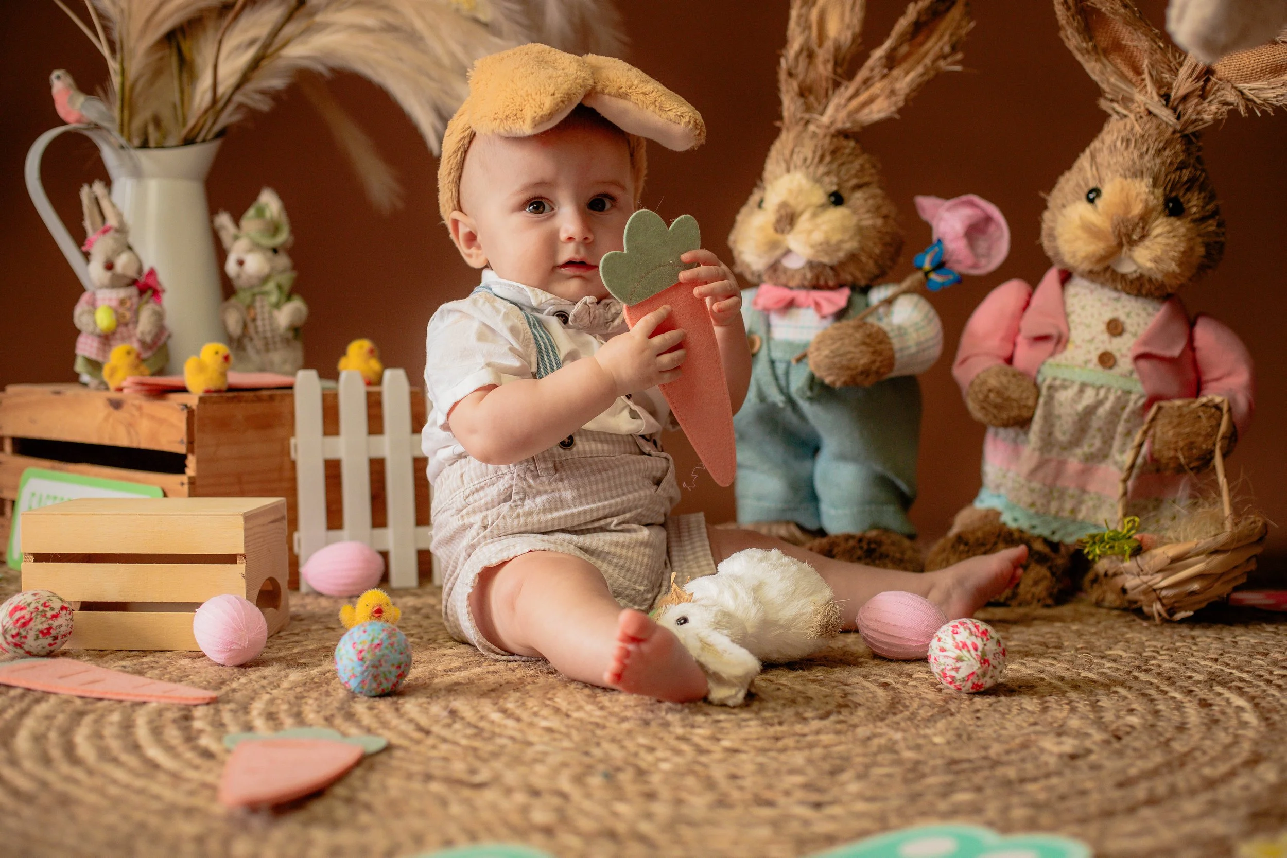 A young child sitting on a woven rug, holding a felt carrot with a green top, surrounded by Easter decorations including plush bunnies, chicks, and cotton balls resembling eggs, with stuffed bunny figures in the background.