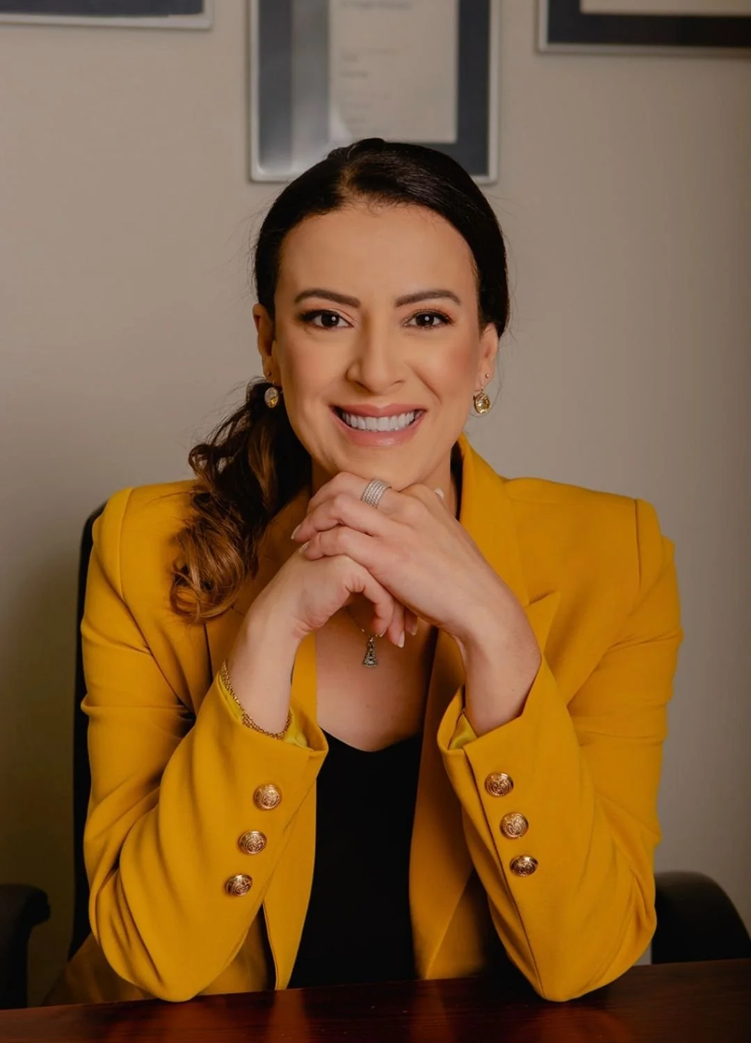 A woman with dark hair in a ponytail, wearing a yellow blazer with gold buttons and jewelry, smiling with hands clasped on a table in front of her.