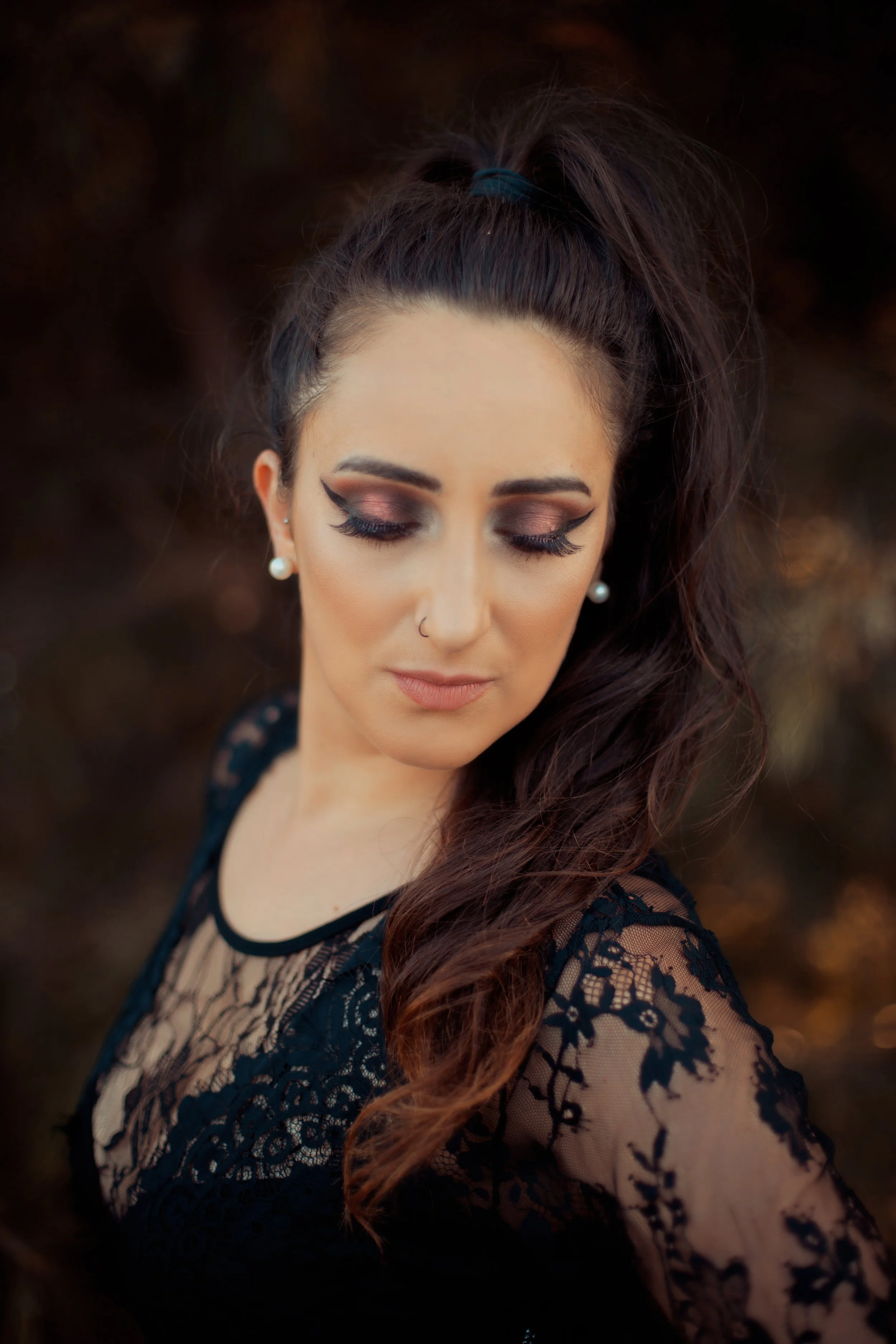 A woman with long dark hair styled in a high ponytail, wearing makeup with cat-eye eyeliner, eyeshadow, and faux lashes, wearing pearl earrings, a nose stud, and a black lace dress with an intricate floral pattern, looking down with a serene expressi
