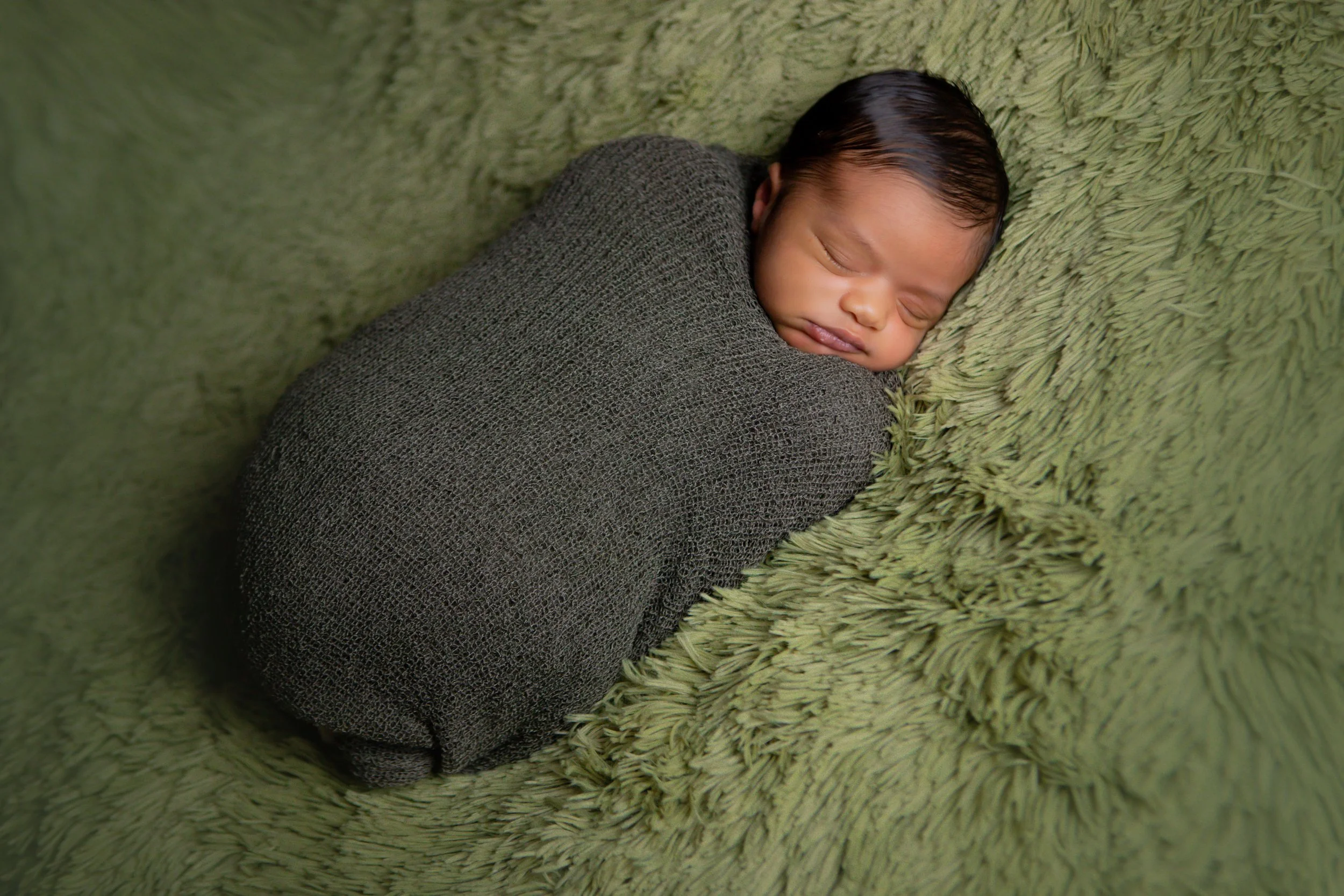 A newborn baby peacefully sleeping wrapped in a dark gray blanket on a textured green blanket.