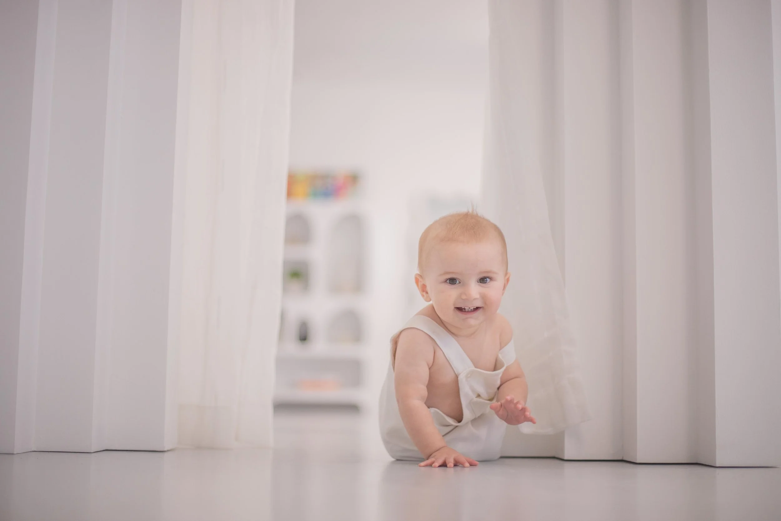 A smiling baby crawling through a doorway with white curtains in a brightly lit room.