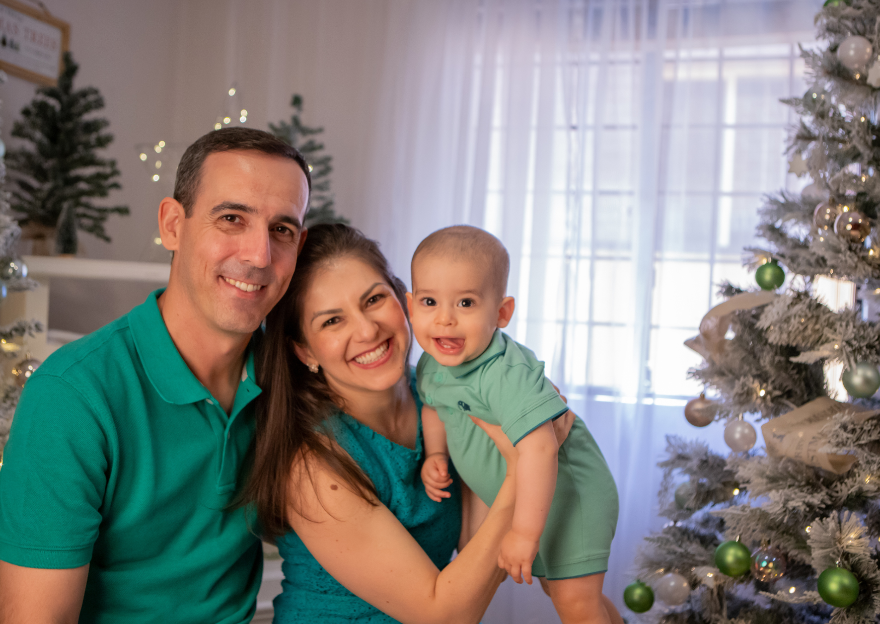 A smiling family of three, including a man, woman, and a young boy, standing in front of a decorated Christmas tree with ornaments and lights, near a window with white curtains.