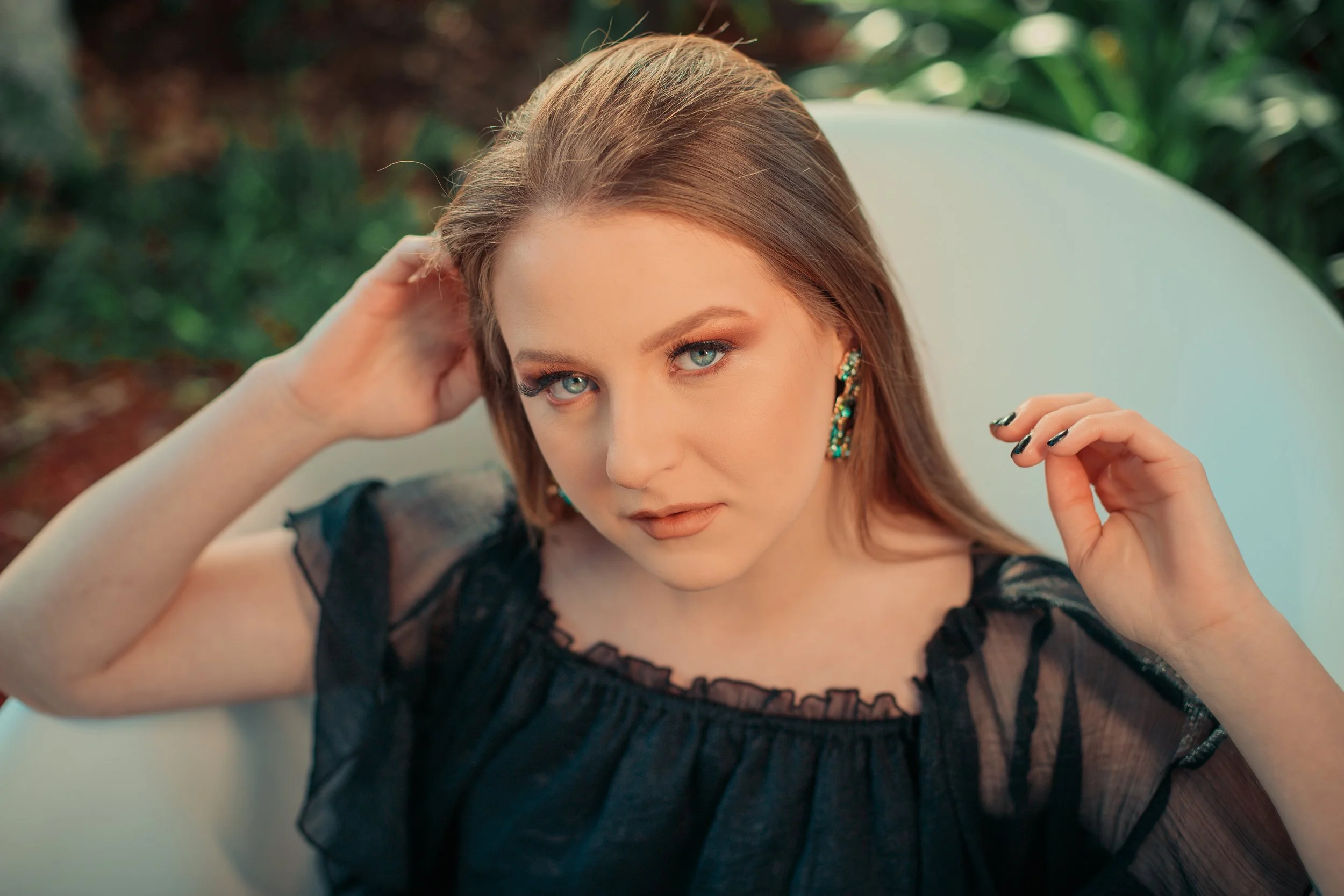 A woman with blue eyes and long light brown hair, wearing black sheer top with lace details and colorful earrings, sitting on a white chair outdoors, looking confidently at the camera.