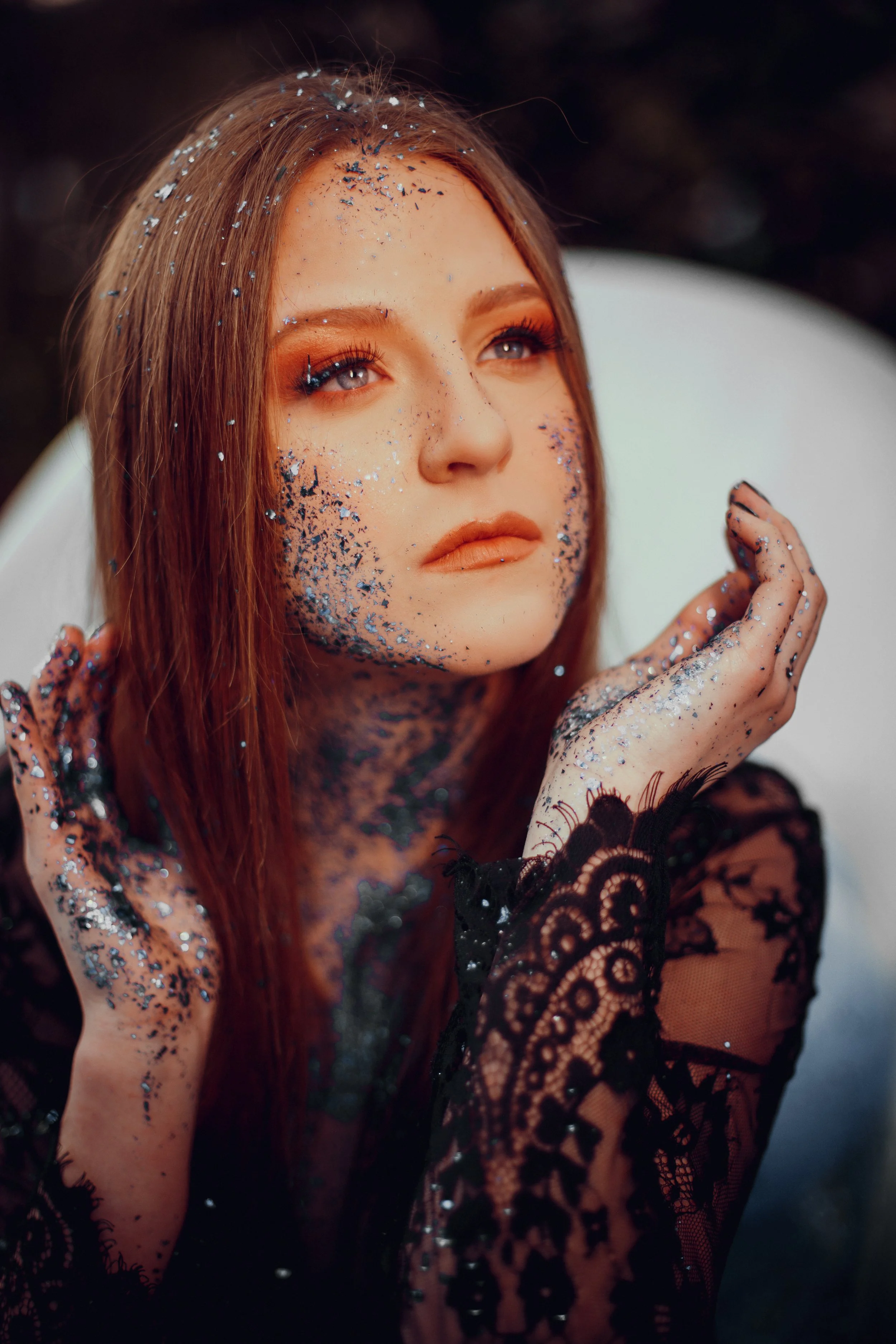 A young woman with long red hair and blue eyes is covered in black and silver glitter, with some glitter on her face, hands, and neck. She is wearing a black lace top and has makeup with orange eyeshadow. She is gazing thoughtfully upward and has her