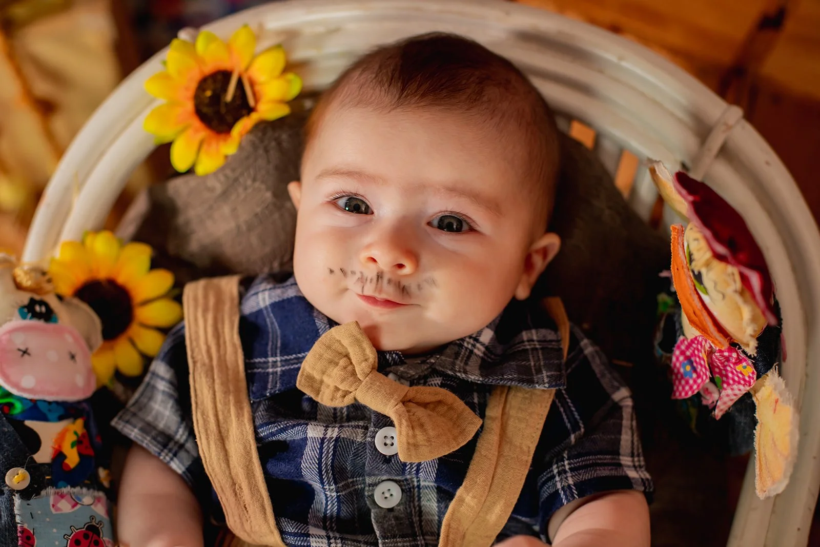 A baby with face paint resembling a mustache and beard, wearing a plaid shirt and a beige bow tie, sitting in a decorated baby seat with sunflower and butterfly decorations.