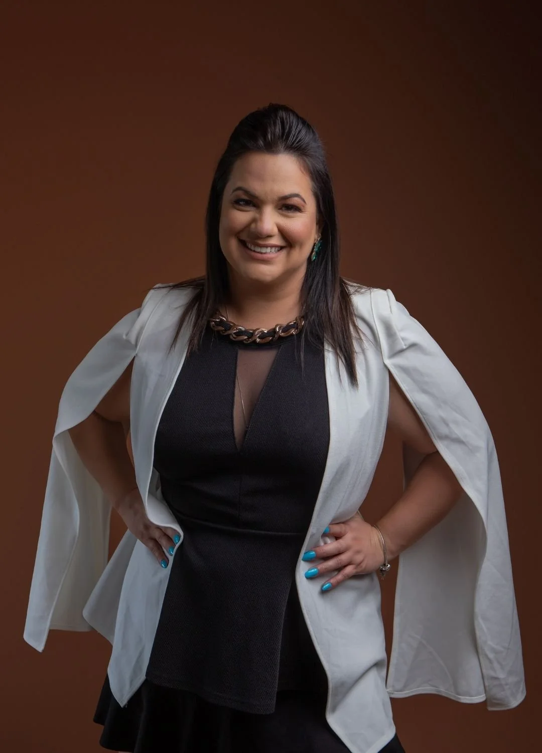 A woman with dark hair smiling, wearing a black dress with a sheer neckline, a white blazer draped over her shoulders, a large chain necklace, earrings, and a bracelet, standing against a brown background with her hands on her hips.