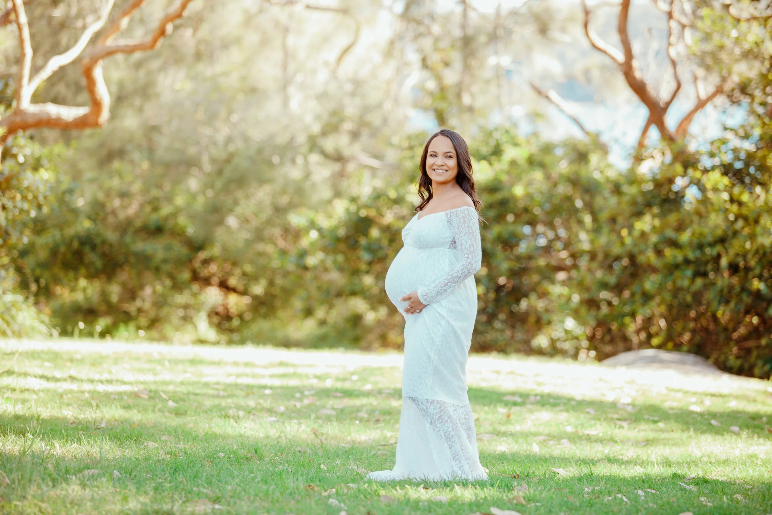 Pregnant woman in a white lace dress standing on grass in a sunlit park.